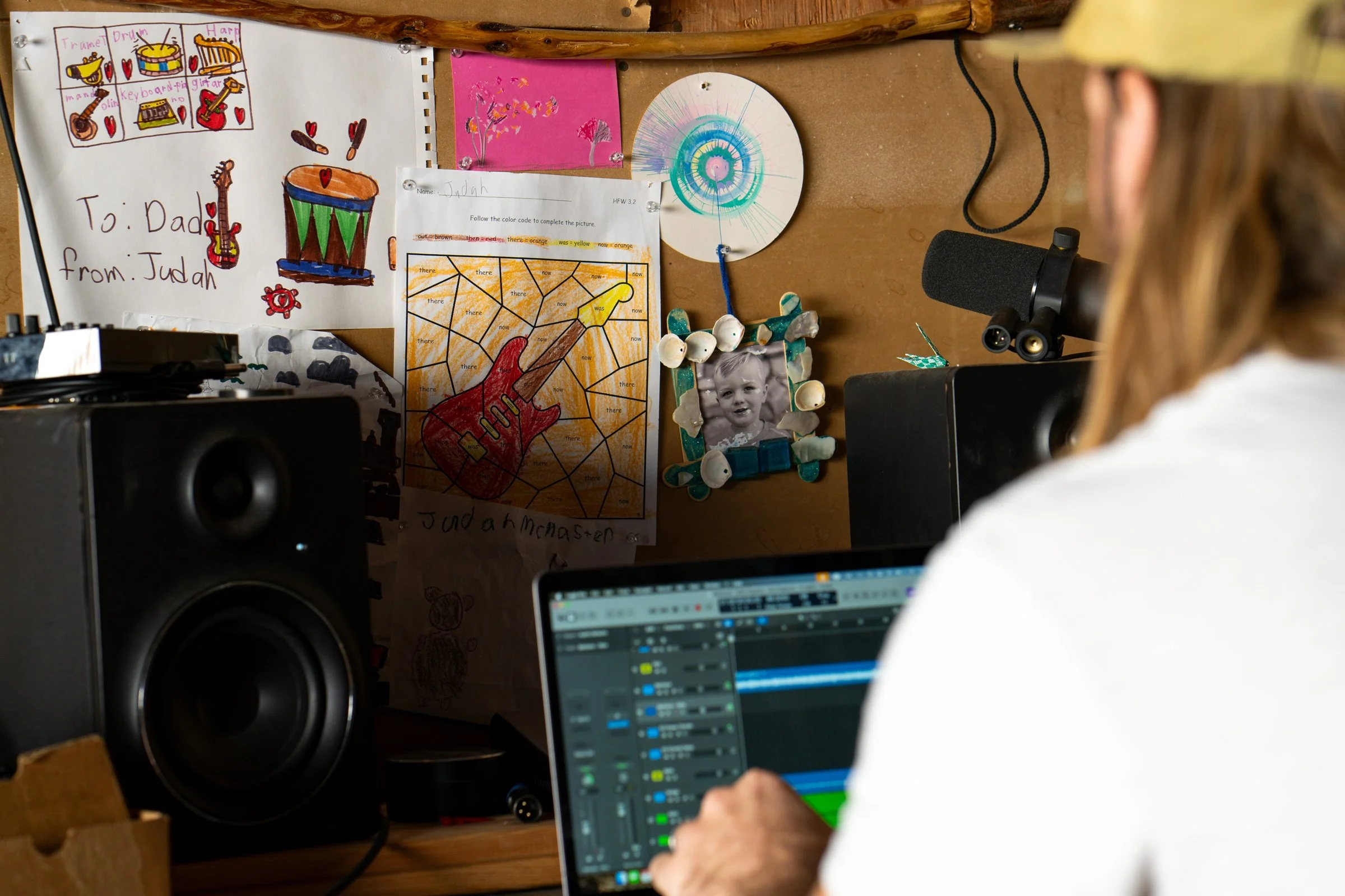 A person working on a music production program on a laptop, with studio speakers and a microphone nearby. The background wall has children's artwork, a framed black-and-white photo of a child, and other decorations.