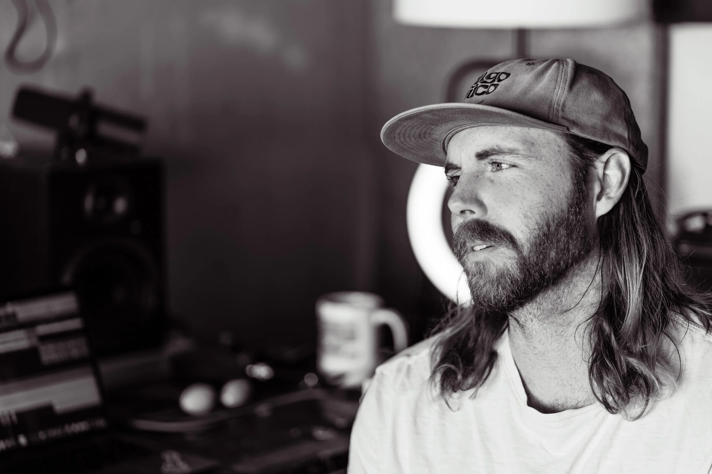 Black and white photo of a man with long hair and a beard, wearing a cap, looking to the right, in a room with sound equipment and a coffee mug in the background.