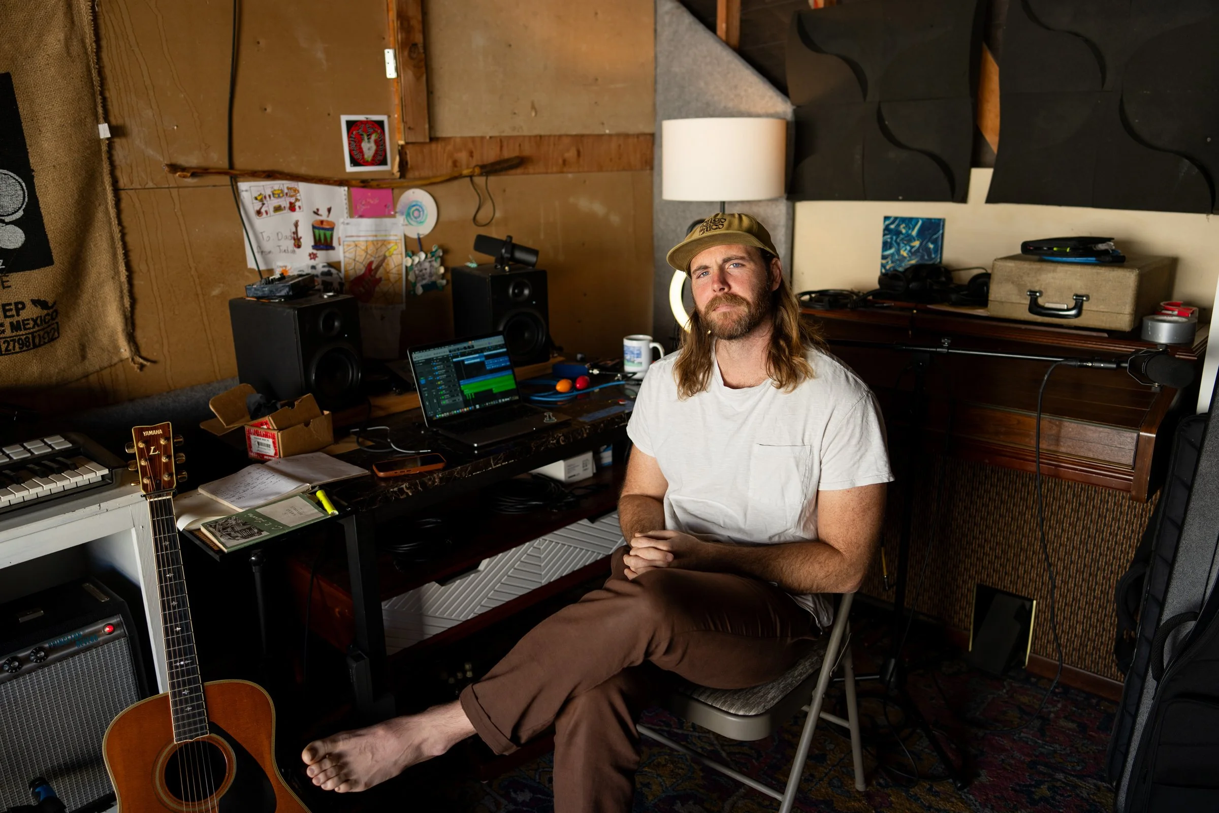A man with shoulder-length hair and a beard, sitting barefoot on a chair in a music studio, wearing a beige cap and white t-shirt.