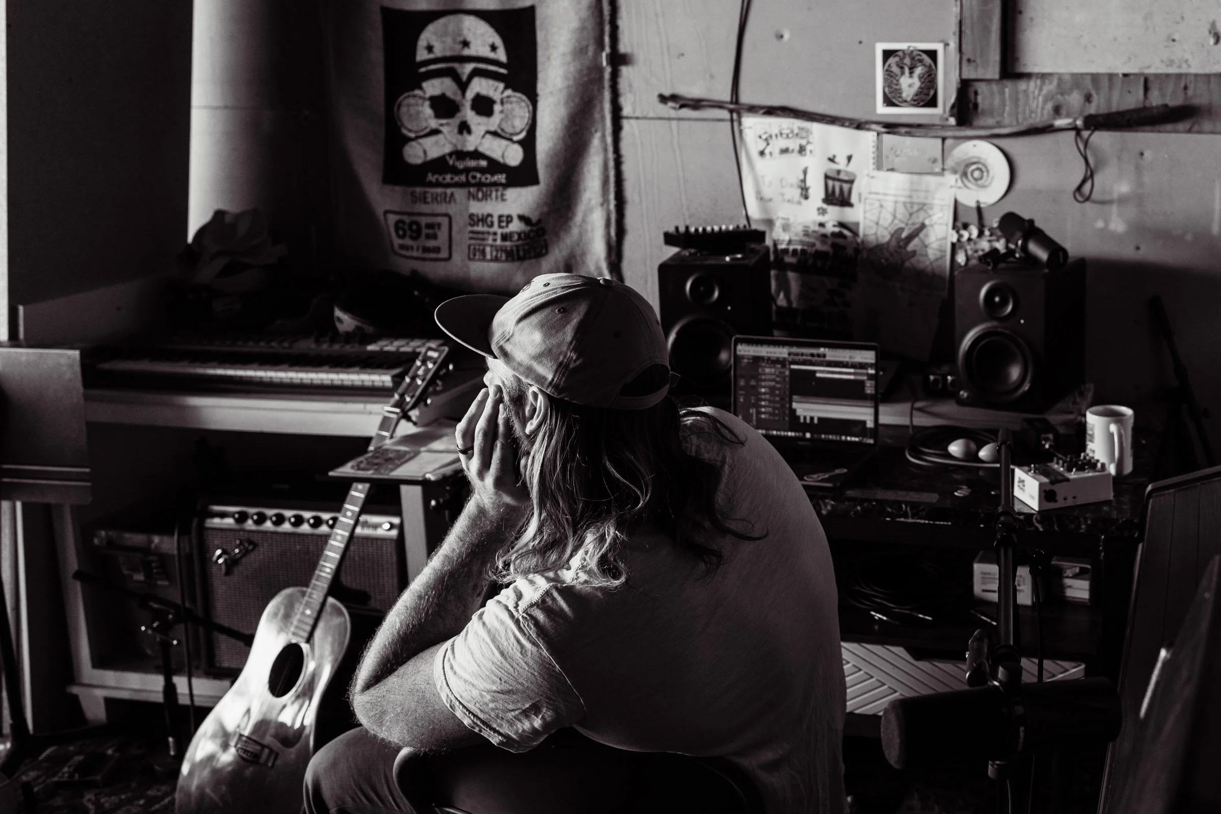 A person with long hair, wearing a cap and T-shirt, sitting in a music studio with their head resting on their hand, surrounded by musical equipment including a guitar, computer, speakers, and posters on the wall.