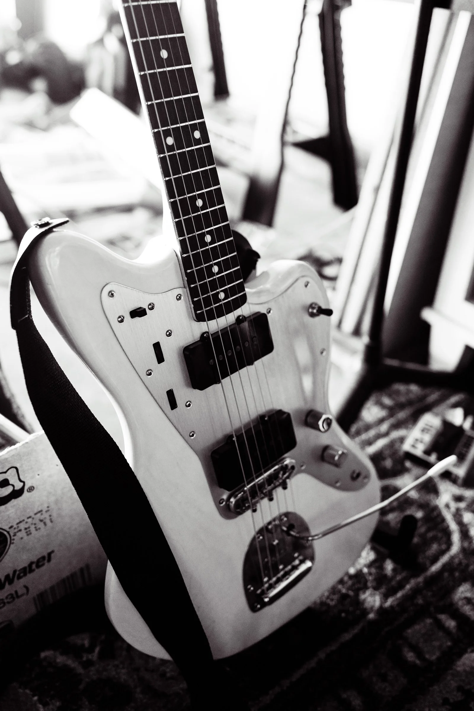 A white electric guitar, with a black strap, standing on a patterned rug, in a room with furniture and various objects in the background.