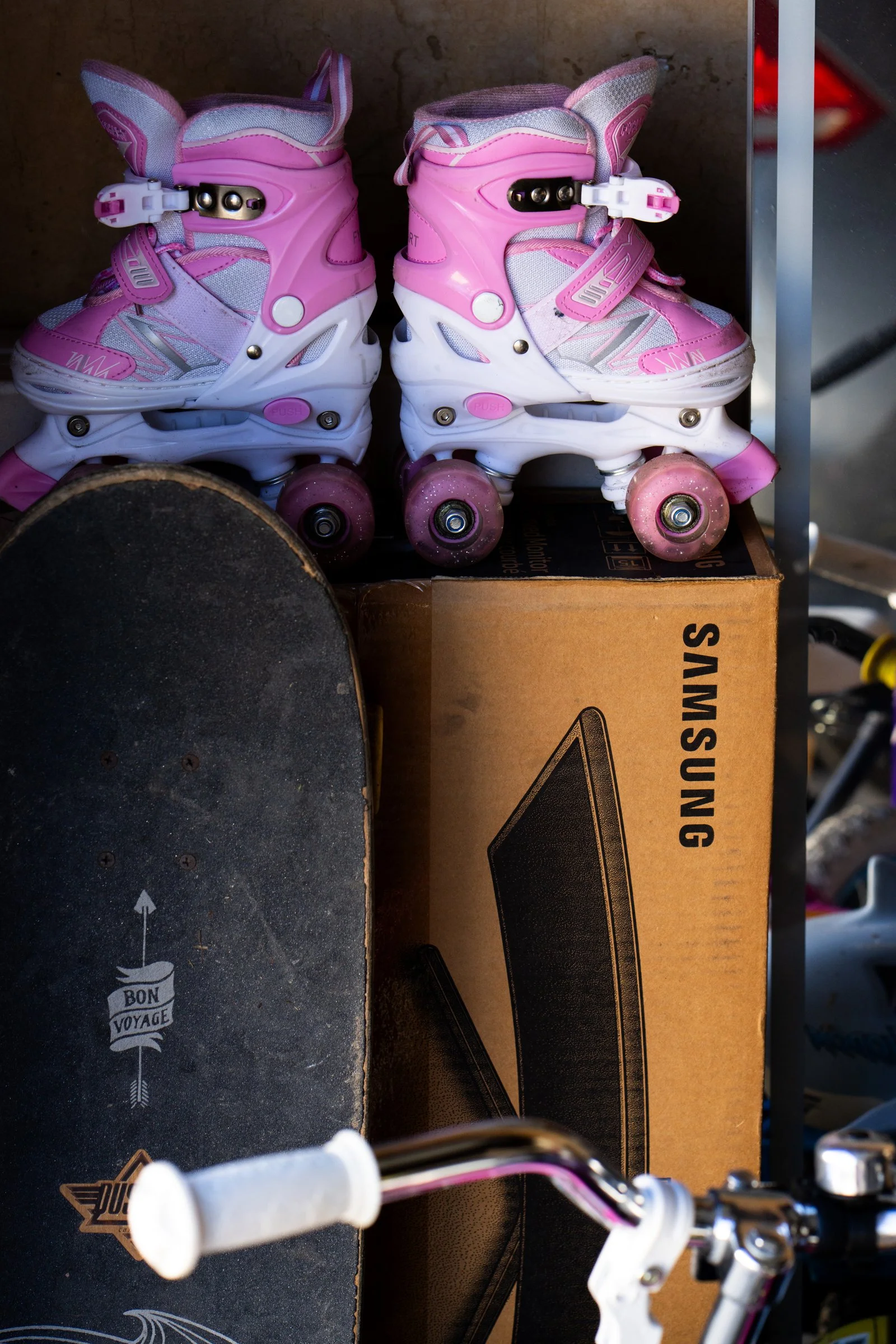 Pair of pink and white rollerblades on top of a cardboard box, with a skateboard and bicycle handlebar in the foreground.