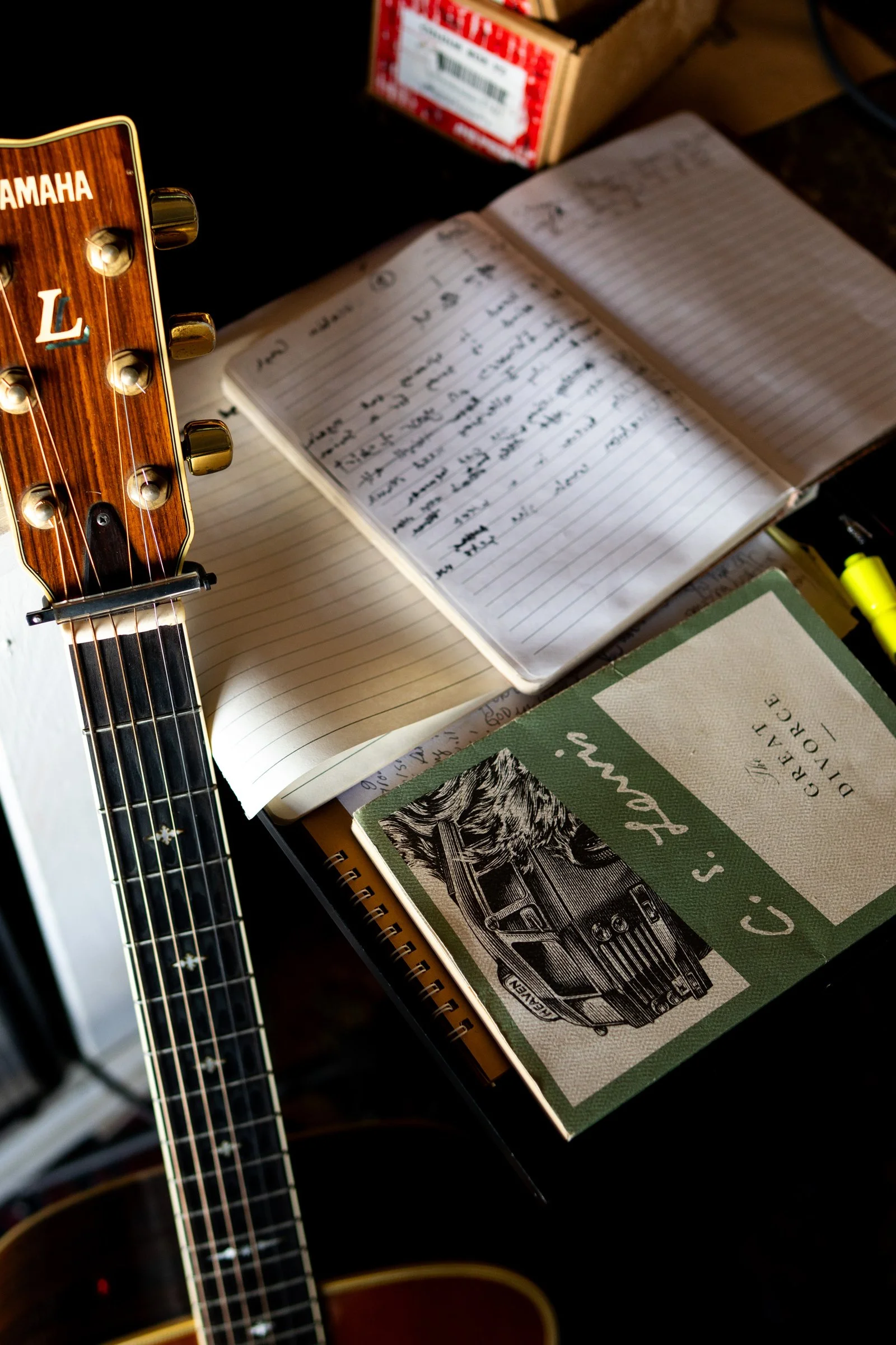 Open notebook with handwritten notes, a vintage C's. Falm book, a Yamaha guitar headstock, and a cardboard box on a dark surface.
