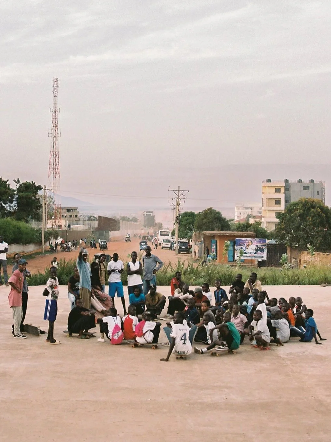 A large group of children and adults gathered outdoors in an open area, some sitting and others standing, with a skater skateboarding on the ground. In the background, there are buildings, trees, and a dusty street with vehicles and people.