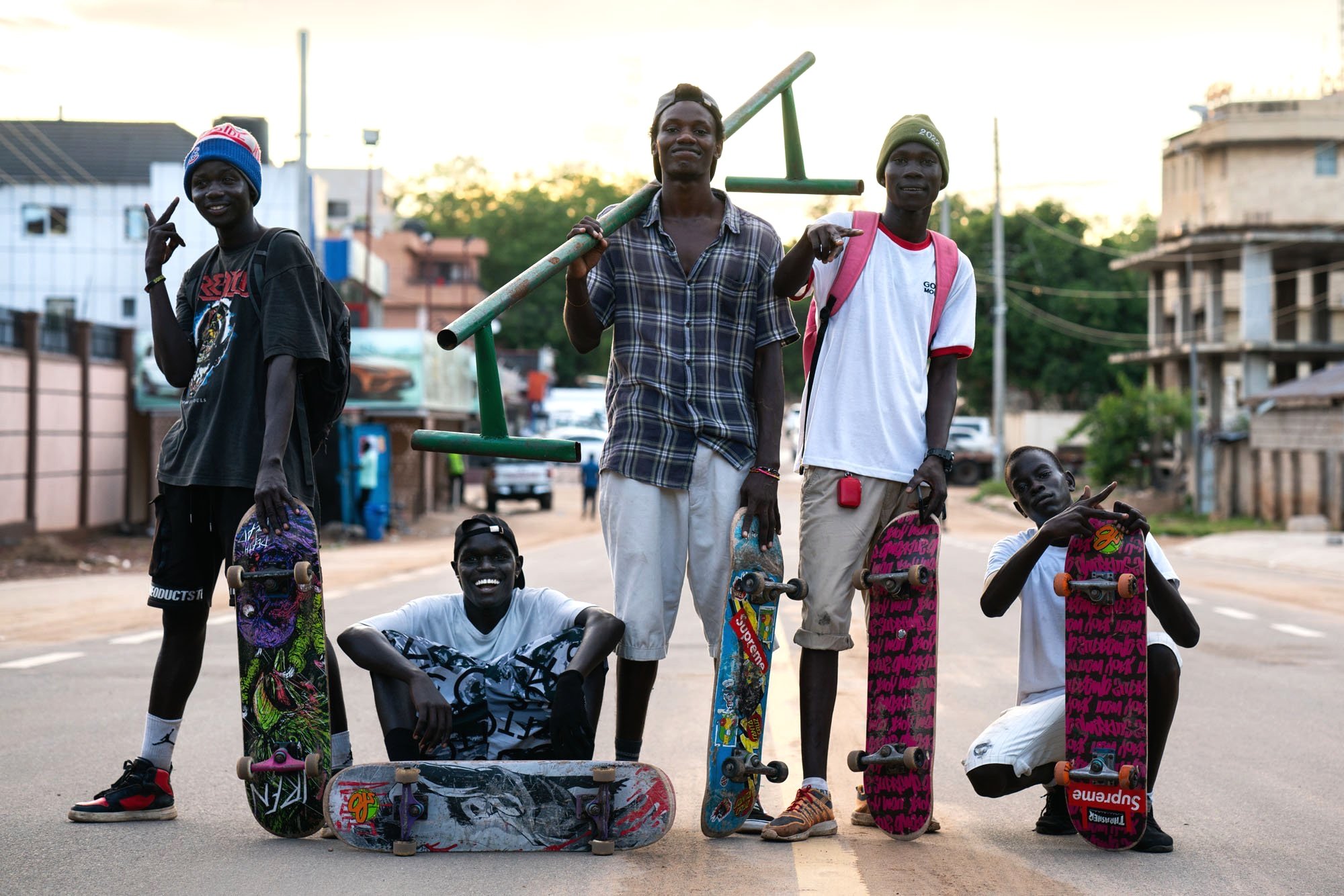 Group of six young boys with skateboards posing on a street in an urban area during the daytime. Some boys are standing, and some are sitting, with skateboards in front of them. They are smiling and making gestures. The background features buildings, trees, and utility poles.