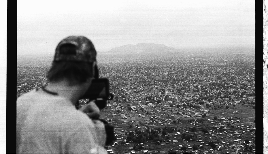 A person aiming a firearm from an elevated position over a densely populated urban area with distant mountains in the background.