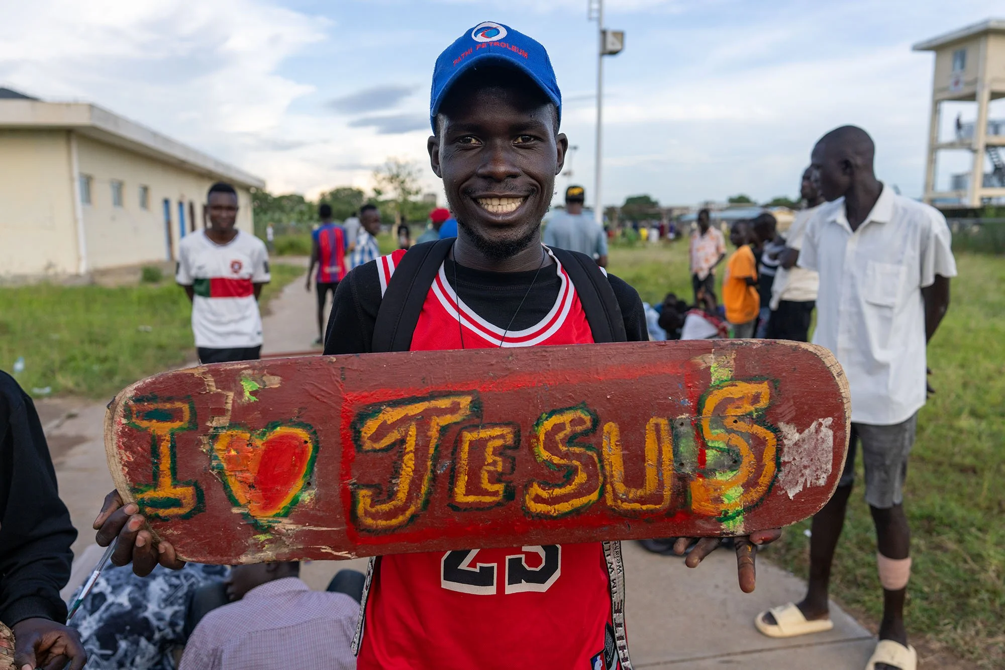 A smiling young man wearing a blue hat and a red sports jersey holds a colorful painted wooden sign that says 'I love TESOS.' He stands outdoors with a group of people in the background, some dressed casually, under a partly cloudy sky.