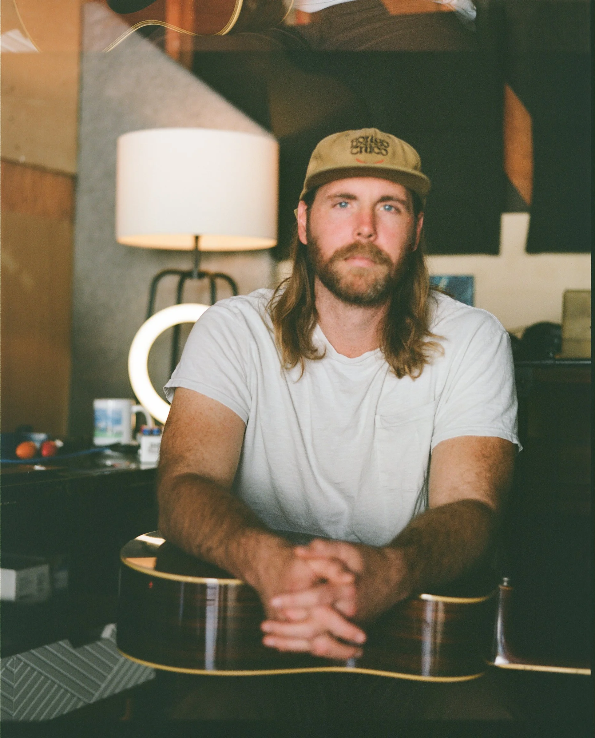 A man with long hair, a beard, and a beige cap sitting at a table with a guitar in front of him. There is a lamp and some objects in the background.