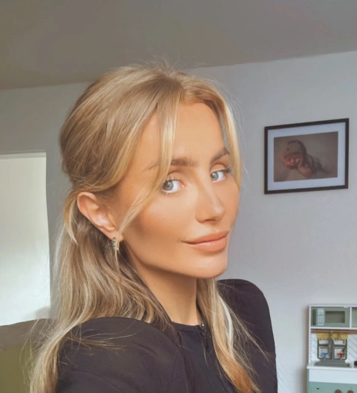 A woman with blonde hair and blue eyes, smiling at the camera, in a room with framed artwork and a toy cash register in the background.