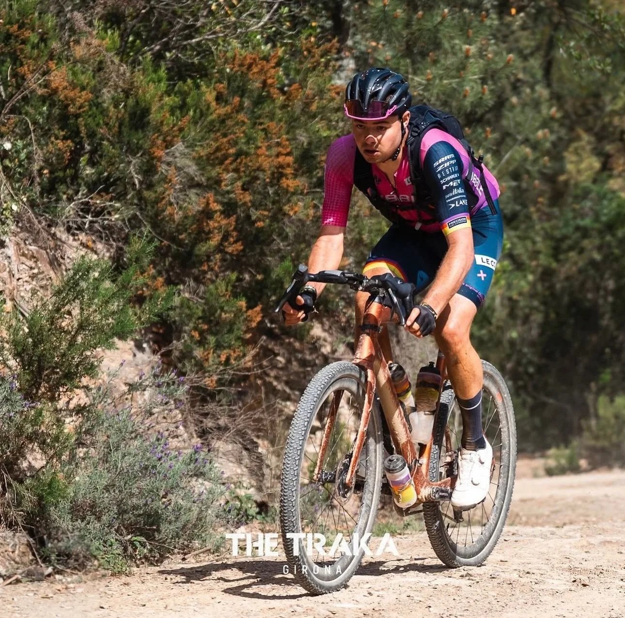 A male cyclist riding a mountain bike on a dirt trail surrounded by green and orange foliage, wearing a black helmet, pink and blue cycling outfit, and black gloves.