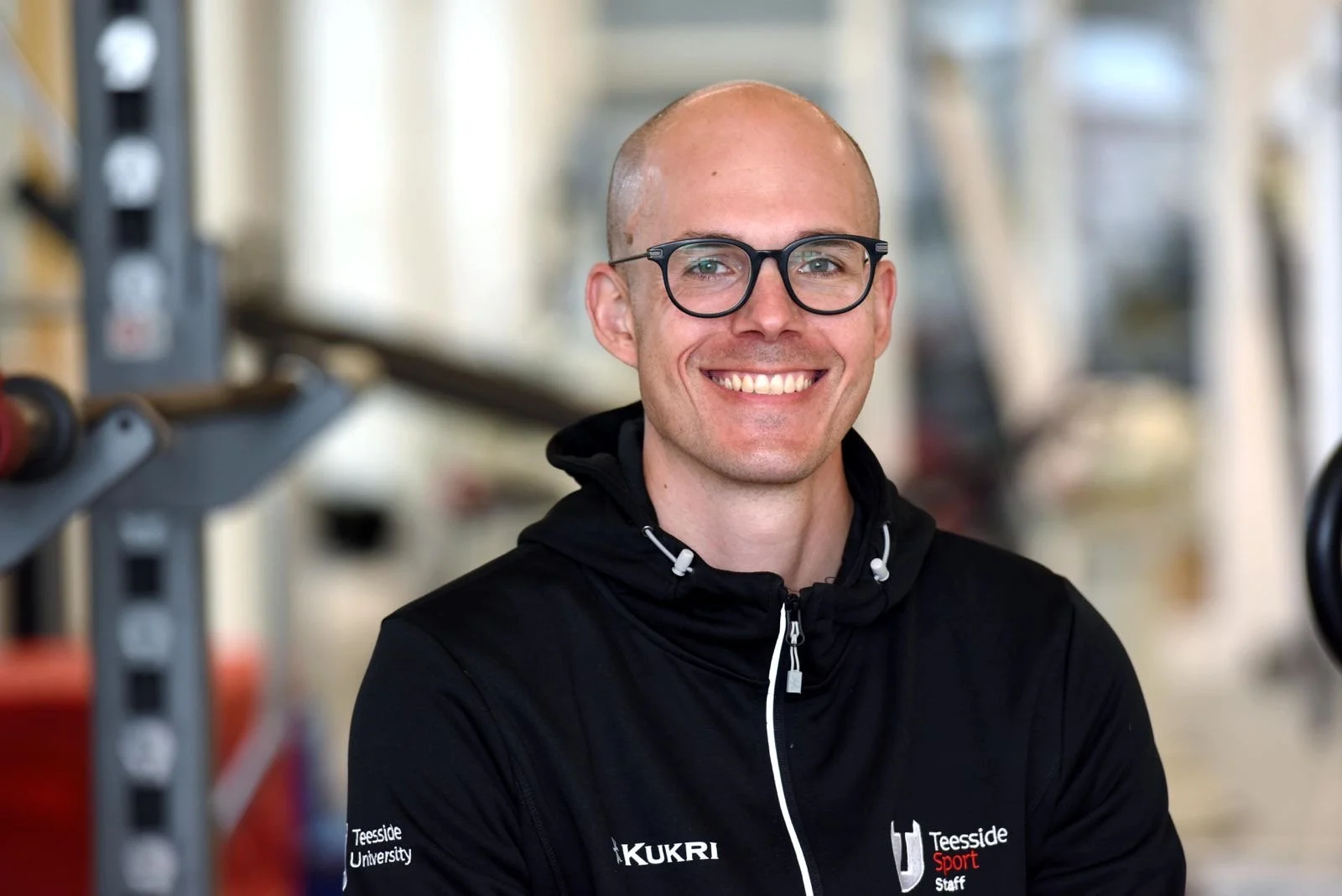 A smiling man wearing glasses and a black sports jacket with 'Teesside University', 'Kukri', and 'Teesside Sport Staff' logos, standing in a gym or fitness facility.