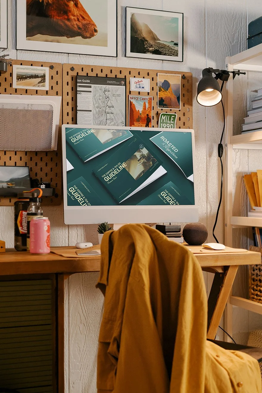A home office setup with a wooden desk, computer screen displaying guides, a yellow jacket draped over a chair, and various items on the desk including a pink beverage can, a black speaker, and some plants. There are framed photographs and a corkboard with travel maps and postcards on the wall behind.