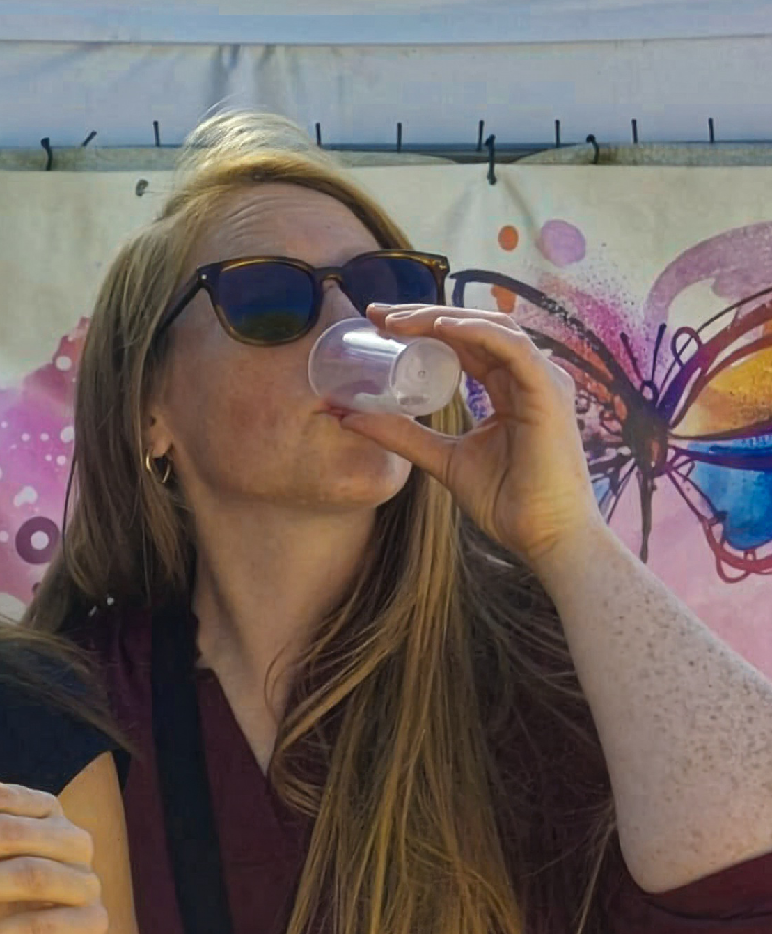A woman wearing sunglasses drinks from a small plastic cup in front of a colorful butterfly mural.