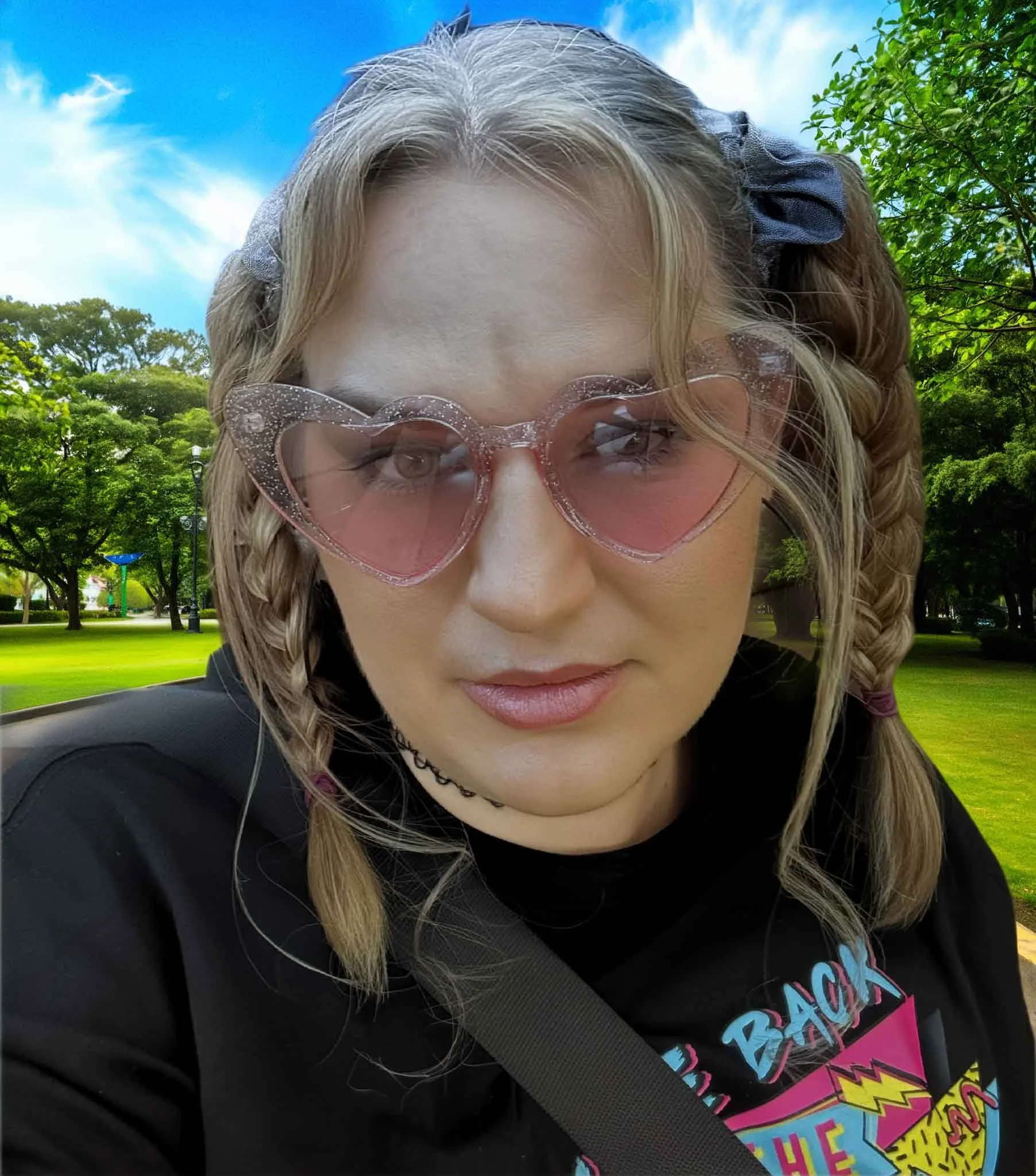 A young woman with blonde hair in two braids wearing pink heart-shaped sunglasses, a black choker, and a black T-shirt with colorful text and graphics, outdoors in a park with green trees and a blue sky.