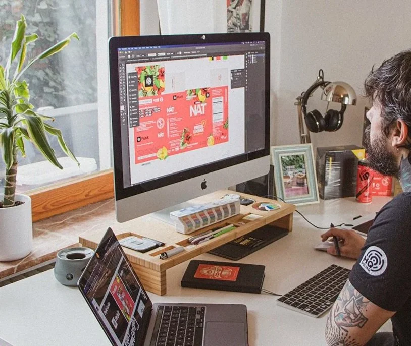 A professional designer working at a desk with a large iMac, a laptop, a small tablet, and various office supplies. The iMac screen displays a design project with a red-themed layout.