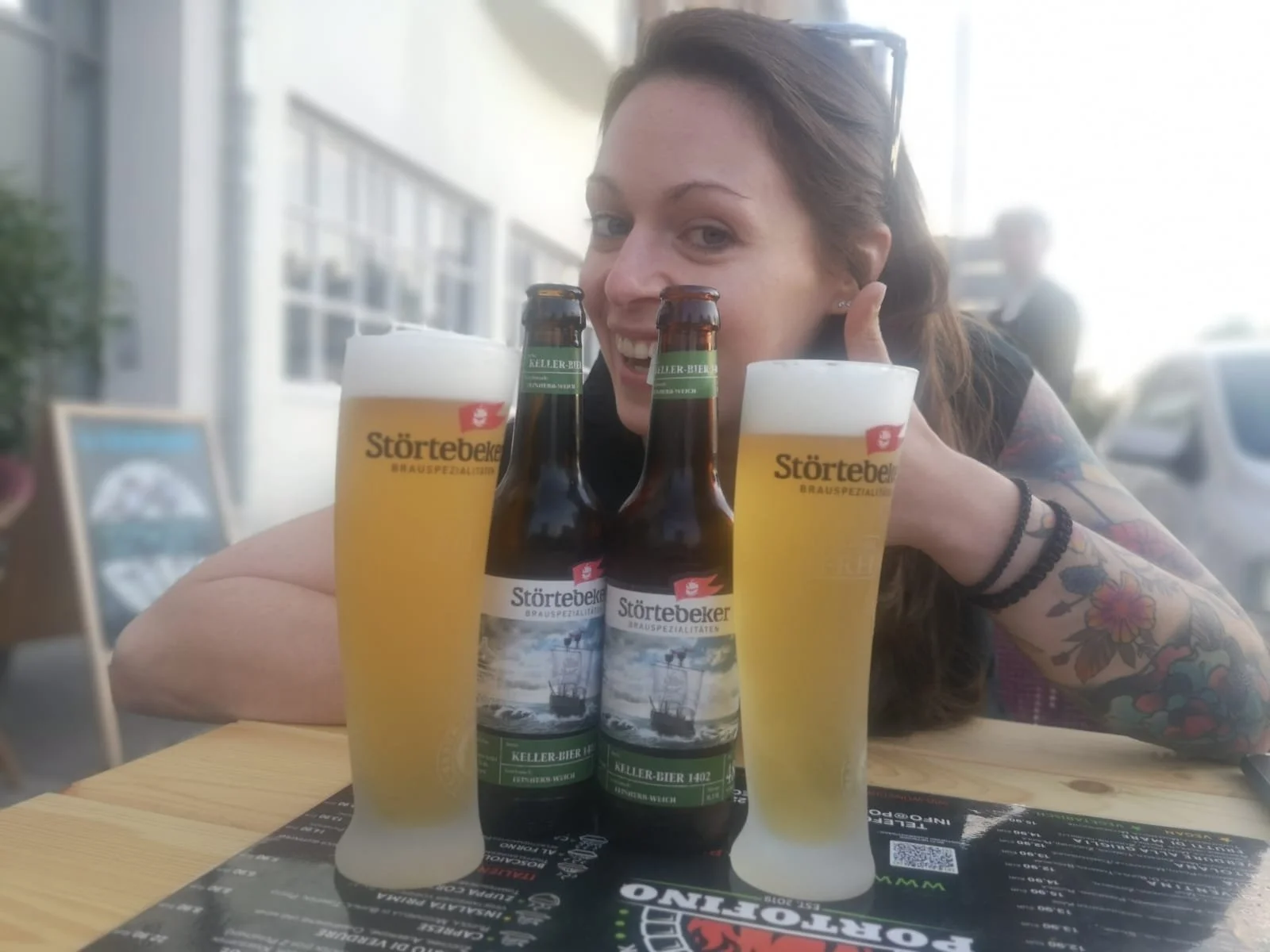 A woman with long hair and tattoos on her arm smiling and giving a thumbs-up, sitting at a table with two glasses of beer and two bottles of Störtebeker beer, outdoors in a sunny setting.