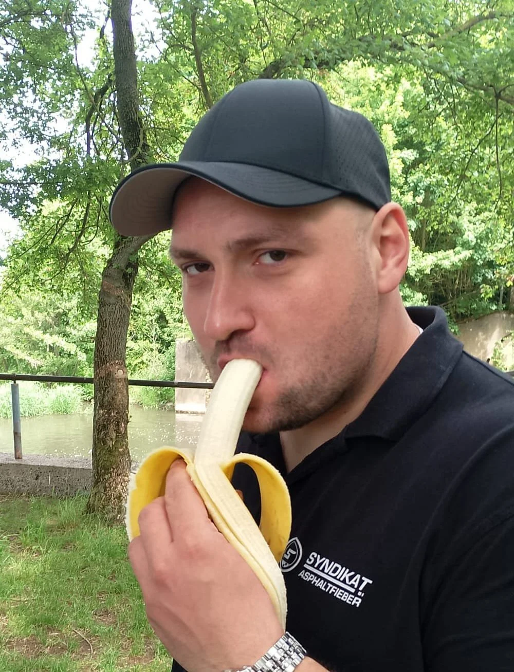 Man in black shirt and black cap eating a peeled banana outdoors near a river and trees