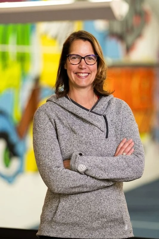 A woman with glasses and shoulder-length brown hair, smiling with arms crossed, standing in a colorful indoor setting that appears to be a store or library.