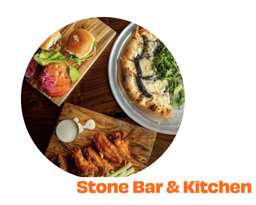 A pizza with cheese and greens on a round metal tray, a wooden board with buffalo wings and a small cup of dipping sauce, and another wooden board with sliders, pickled vegetables, and a side of pickles, all on a wooden table at Stone Bar & Kitchen.