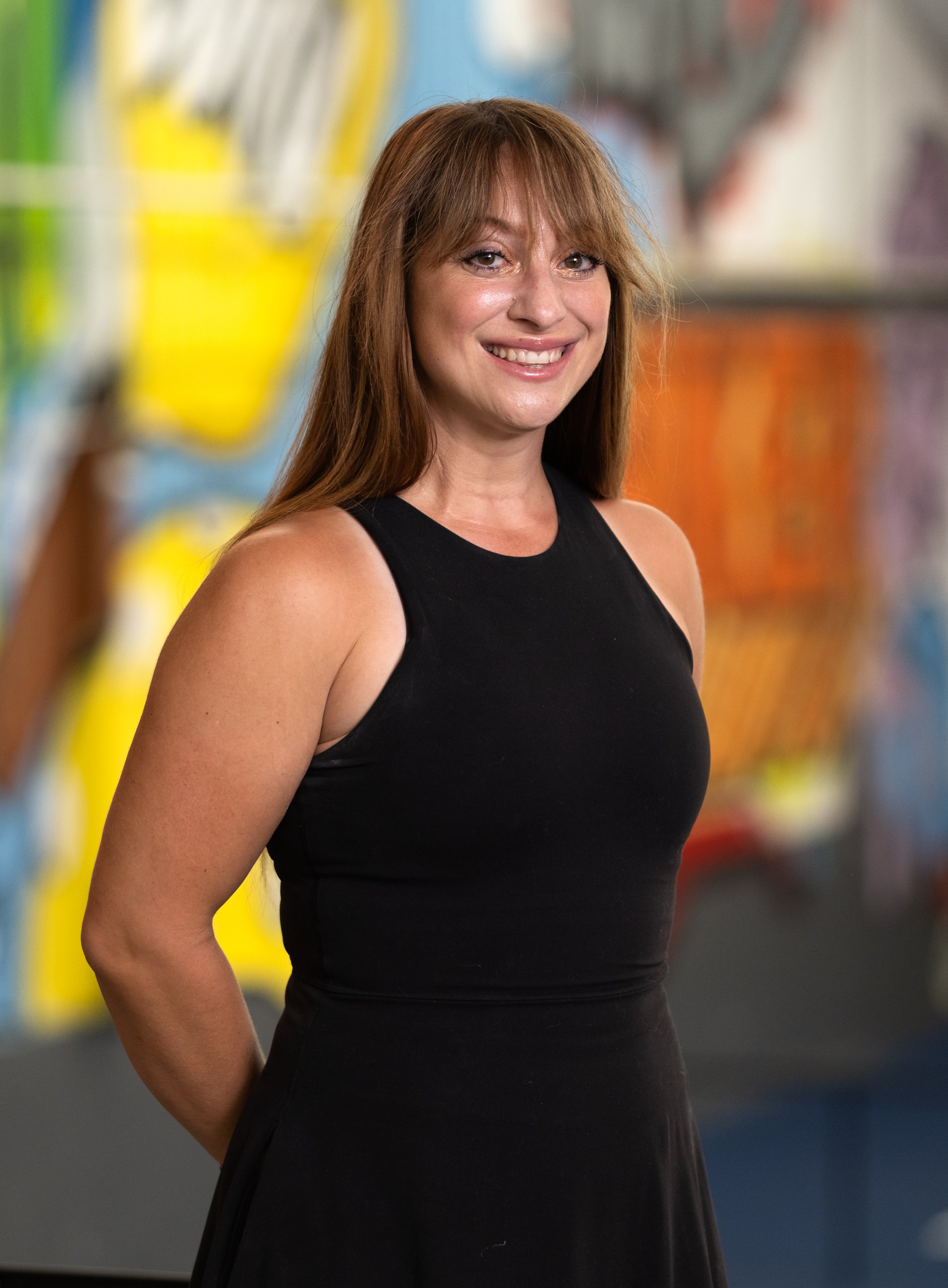 A smiling woman with long reddish-brown hair wearing a sleeveless white top, standing in front of a colorful graffiti wall.