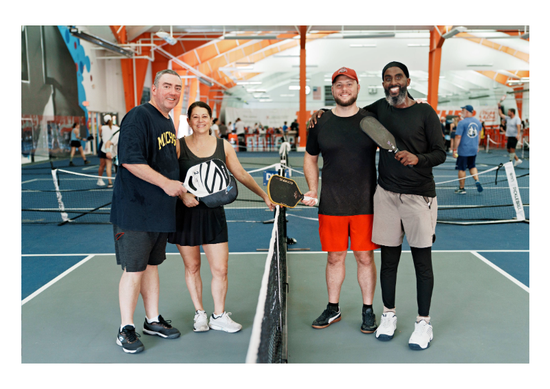 Four people standing on an indoor pickleball court holding paddles, smiling at the camera, with other players and courts visible in the background.
