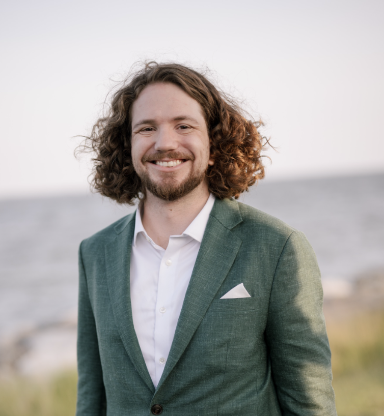 Black and white portrait of a young man with dark hair and beard, wearing a suit and tie, looking directly at the camera against a plain background.