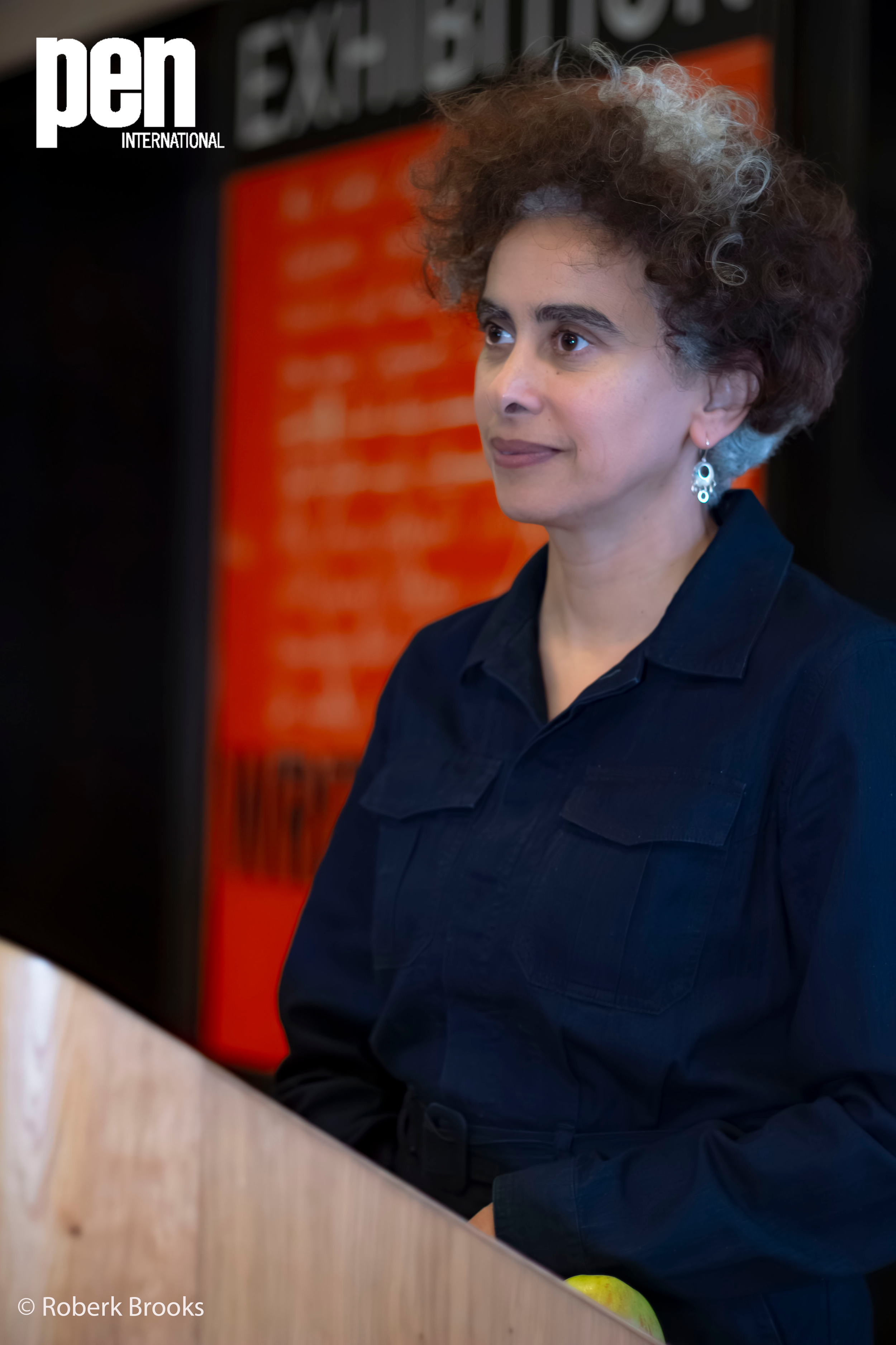 A woman with curly brown and gray hair wearing a dark blue jacket stands near a wooden counter, with a blurred orange sign in the background. She's looking to her right with a neutral expression.