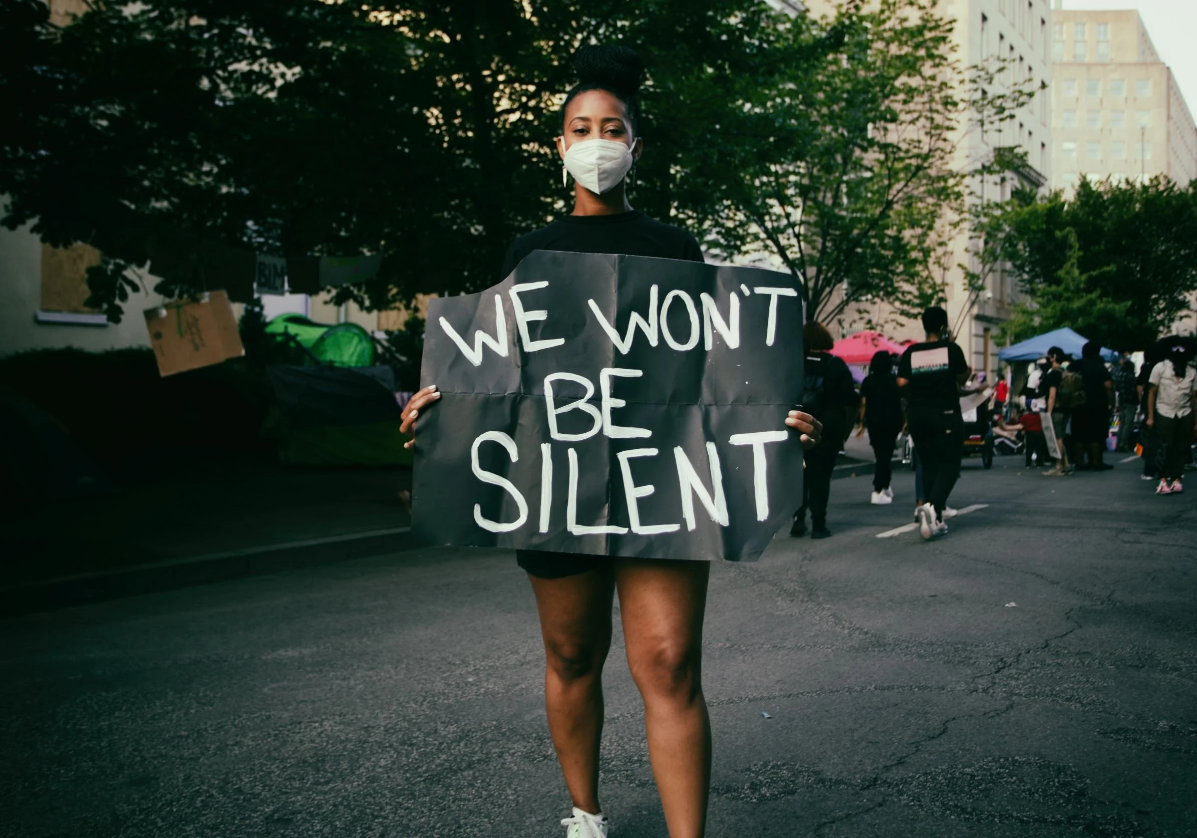Woman wearing a face mask holding a sign that reads "We Won't Be Silent" during a protest on a city street, with other protesters and tents visible in the background.