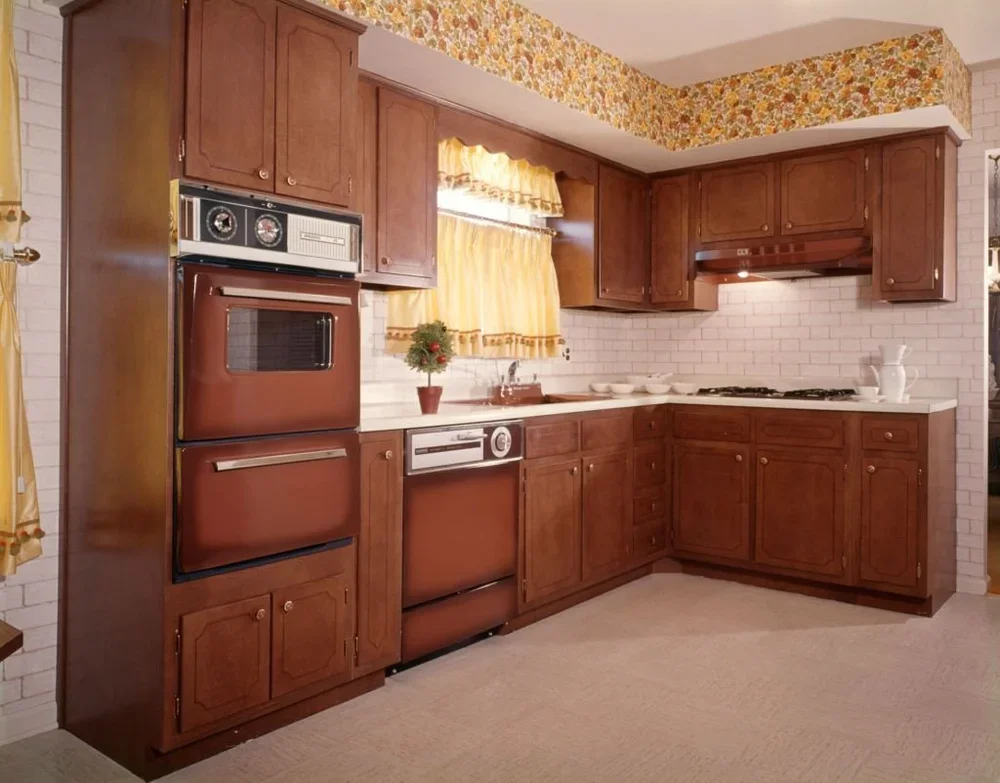 Vintage kitchen with wooden cabinets, a built-in oven, a countertop, a small window with yellow curtains, and a gas stove with white ceramic pitcher.