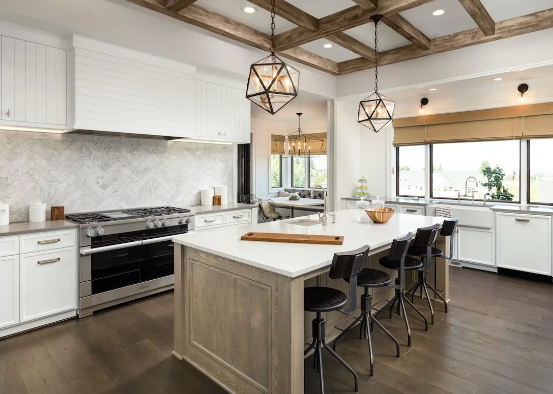 Modern kitchen with white cabinets, marble backsplash, fireplace, large island with black chairs, wood-beamed ceiling, pendant lights, large windows with beige shades, and a small dining area in the background.