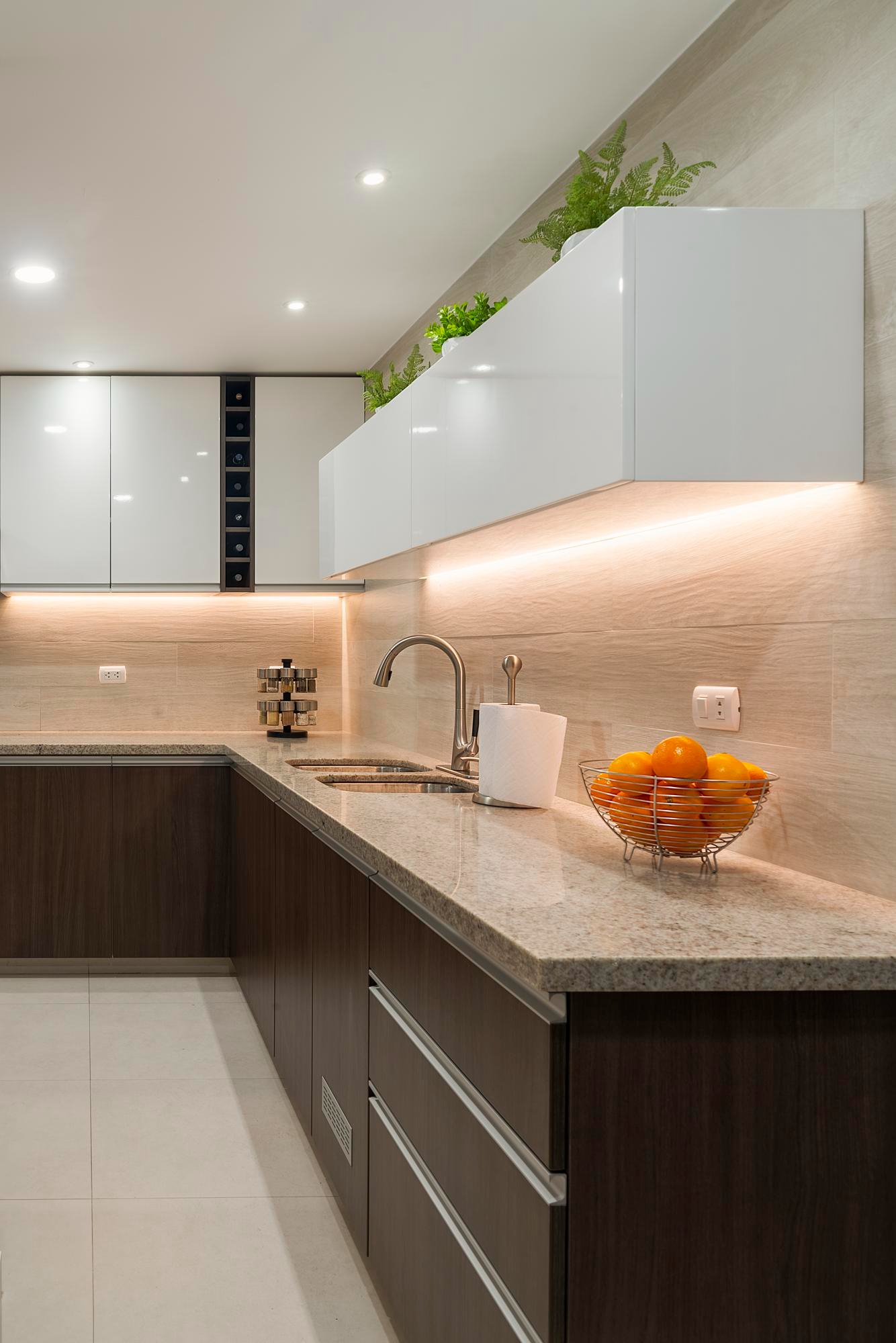 Modern kitchen with beige granite counter, dark wood lower cabinets, white wall cabinets, potted plants, bowl of oranges, paper towel holder, and under-cabinet lighting.