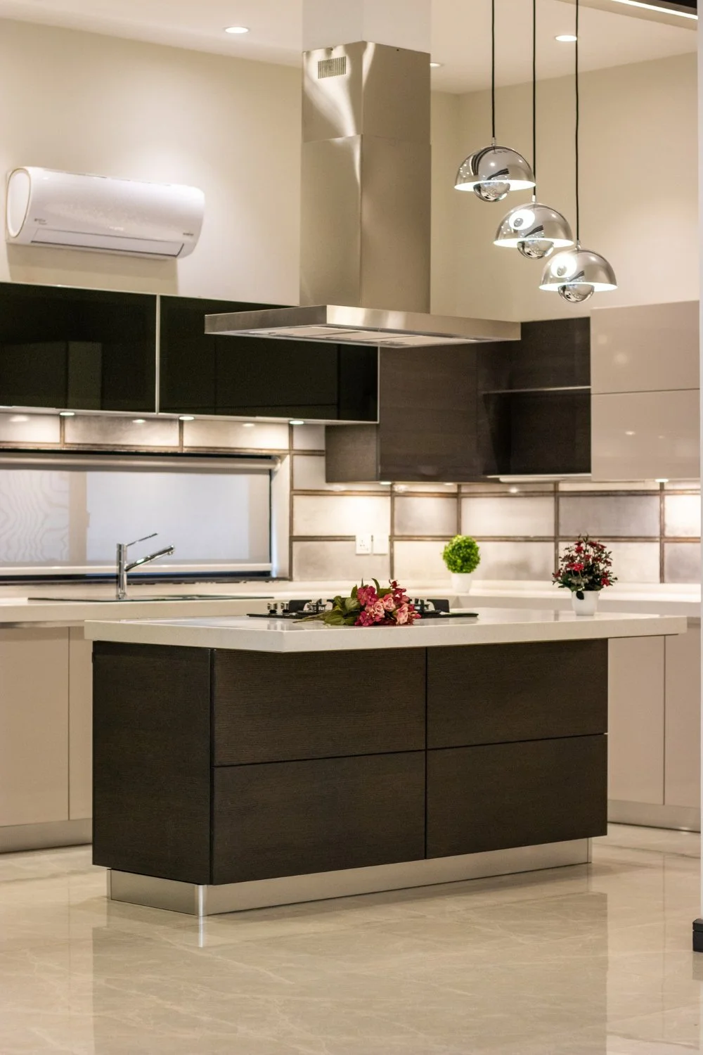 Modern kitchen with dark and light cabinetry, a kitchen island with a flower arrangement, pendant lights, a window, and potted plants on the counter.