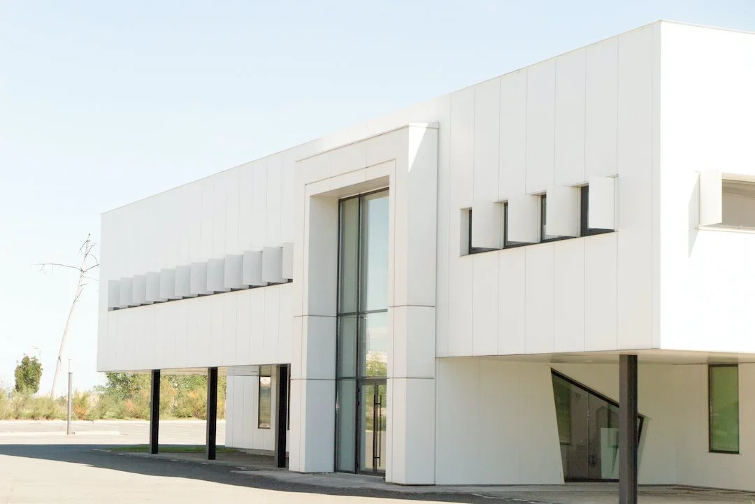 Modern white building with large glass windows and black support columns, surrounded by an empty parking lot.