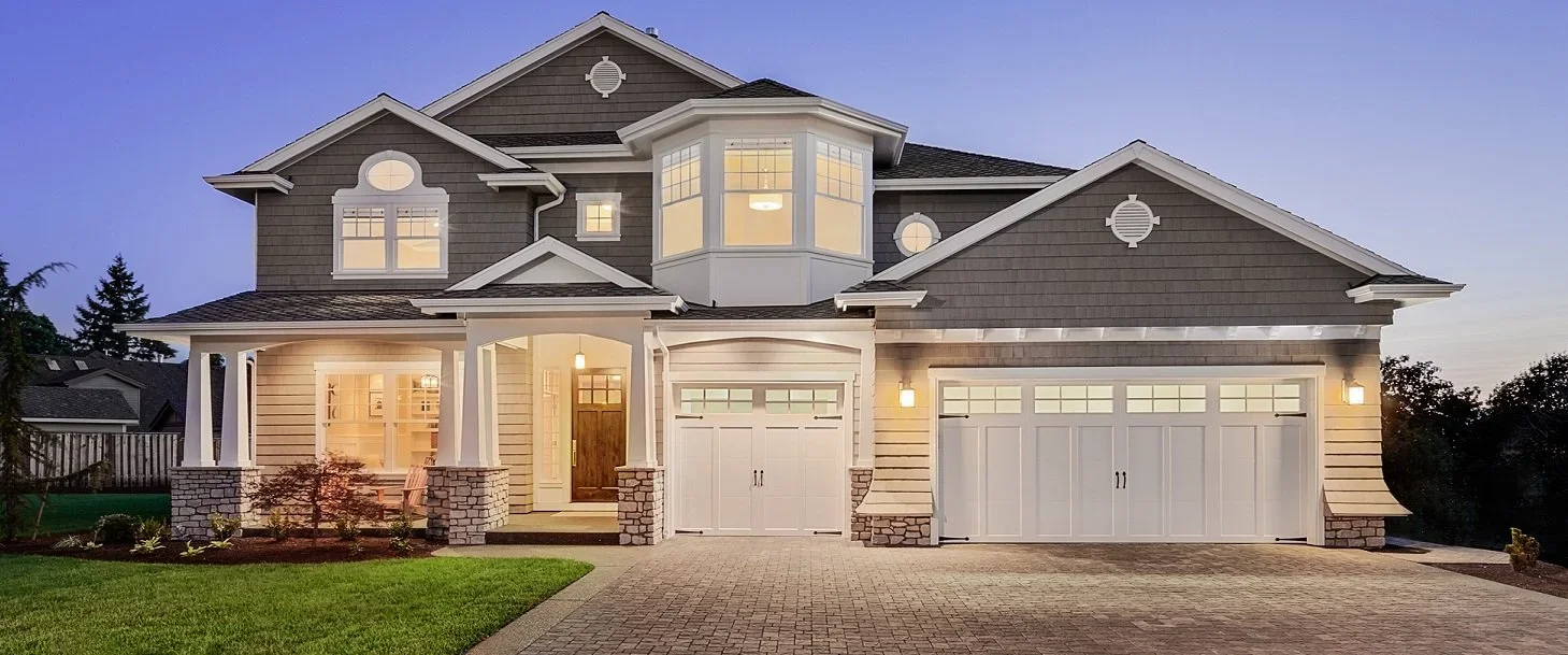 A modern two-story house with gray siding and white trim, illuminated at dusk, featuring a landscaped front yard, a paved driveway, and multiple garage doors.