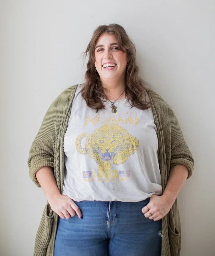 Woman with wavy brown hair wearing a white graphic T-shirt with a lion and the words 'Queen', a green cardigan, and blue jeans, standing in front of a plain wall, smiling.