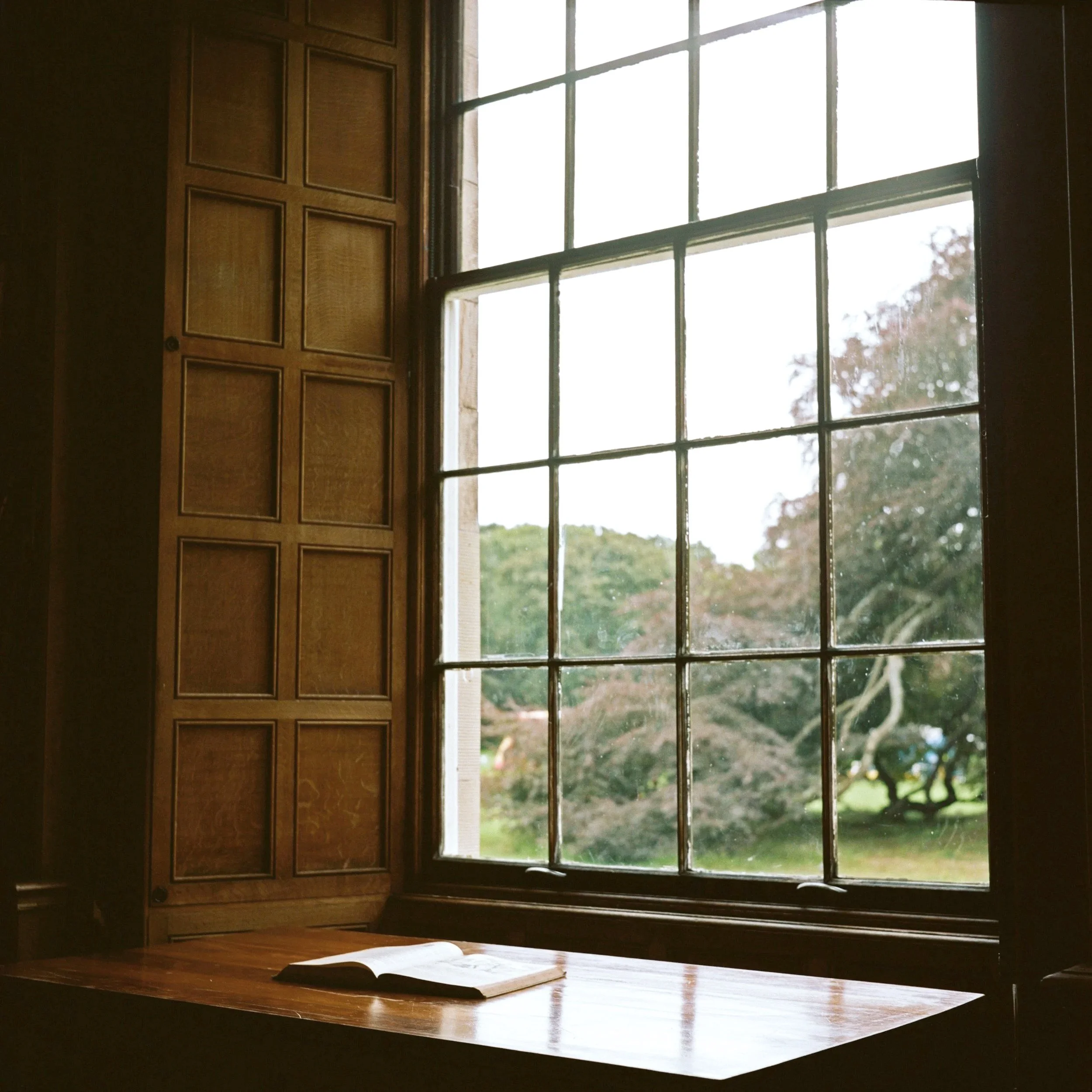 A wooden window with a grid of small panes, open shutters on the side, and a wooden desk with an open book underneath the window.