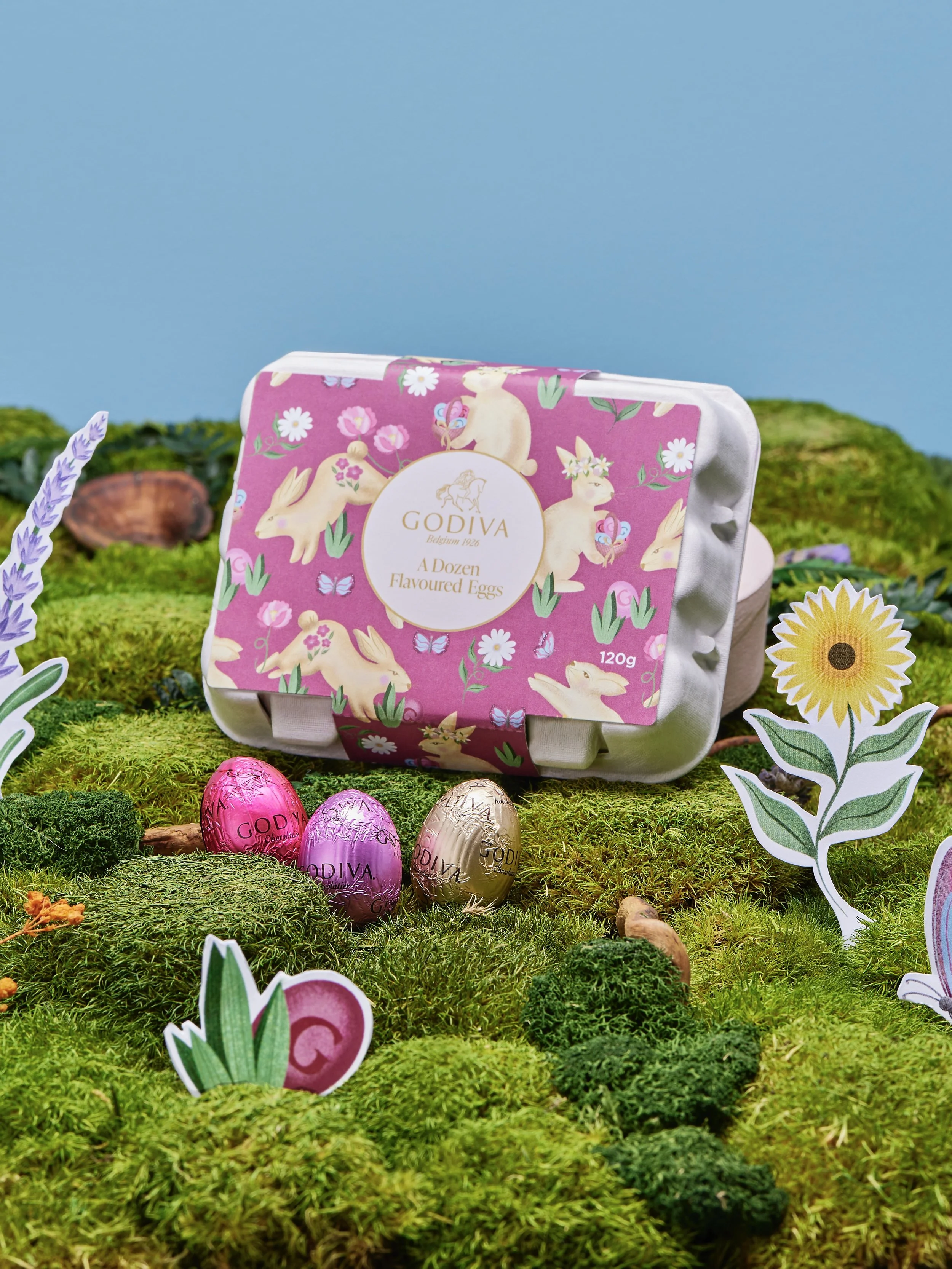 A pink and white package of Godiva flavored eggs with bunny designs, placed on a green moss-like surface decorated with paper flowers and leaves, against a blue sky background.