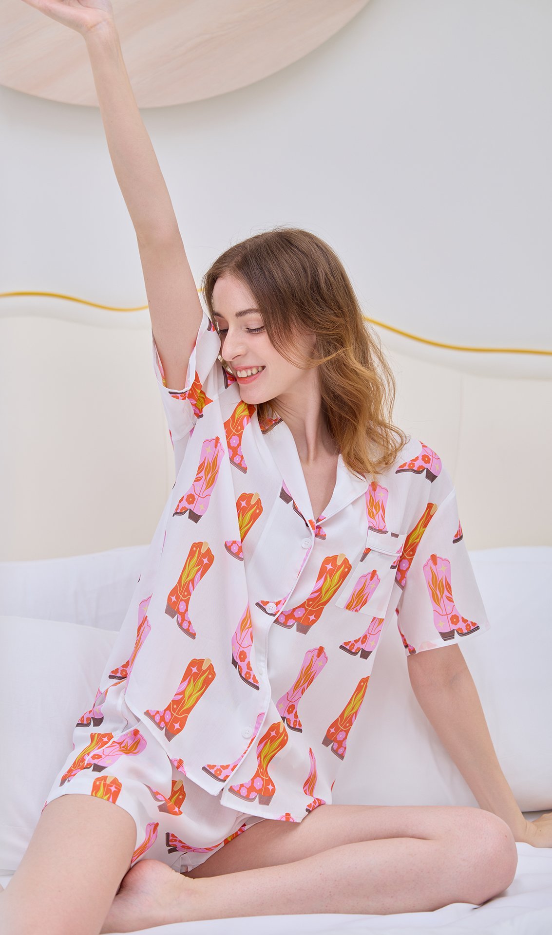 A young woman with wavy brown hair sitting on a bed, wearing a white pajama shirt and shorts decorated with pink, orange, and yellow boots. She is smiling with her eyes closed and has her right arm raised, stretching.