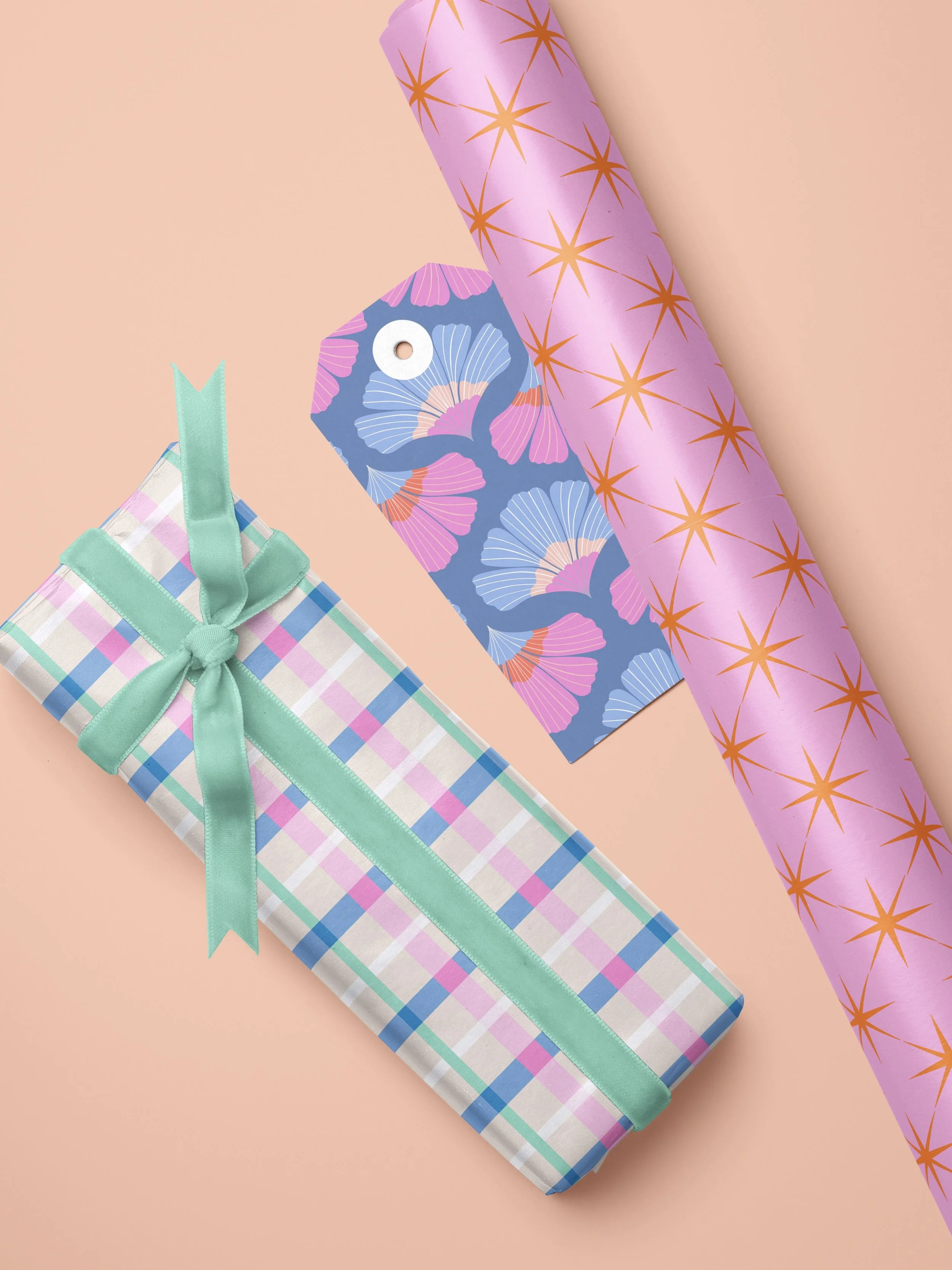 A pastel pink surface with gift-wrapped presents and wrapping paper. The presents include a rectangular gift with pastel plaid paper tied with a mint green ribbon and bow, and a rectangular gift tag with a blue background and pink and purple ginkgo l
