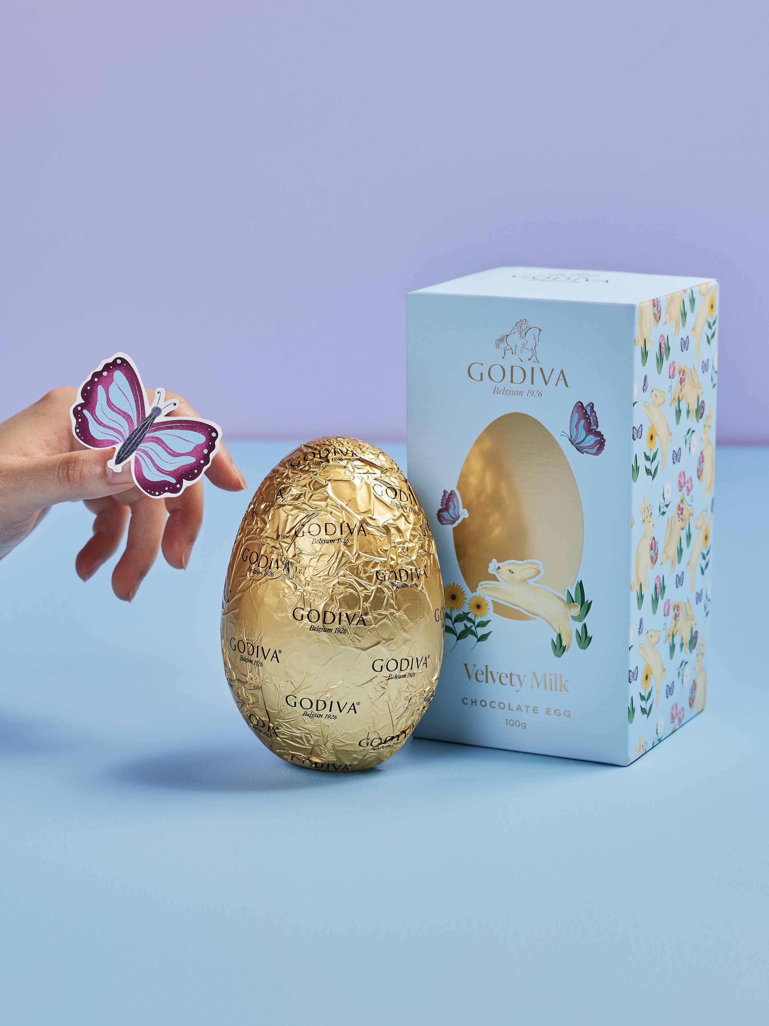 Gold foil wrapped chocolate egg with Godiva branding, next to a pastel blue box with bunny and floral illustrations, and a hand holding a pink and purple butterfly sticker.