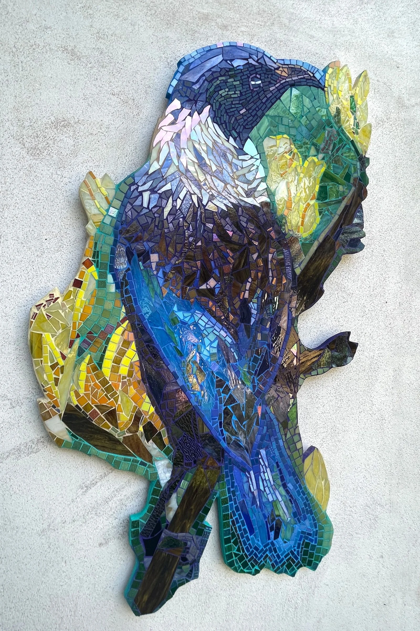 Mosaic art of a Tui bird in profile with its head turned to the right, made from various small, colored glass pieces on a light gray textured wall.