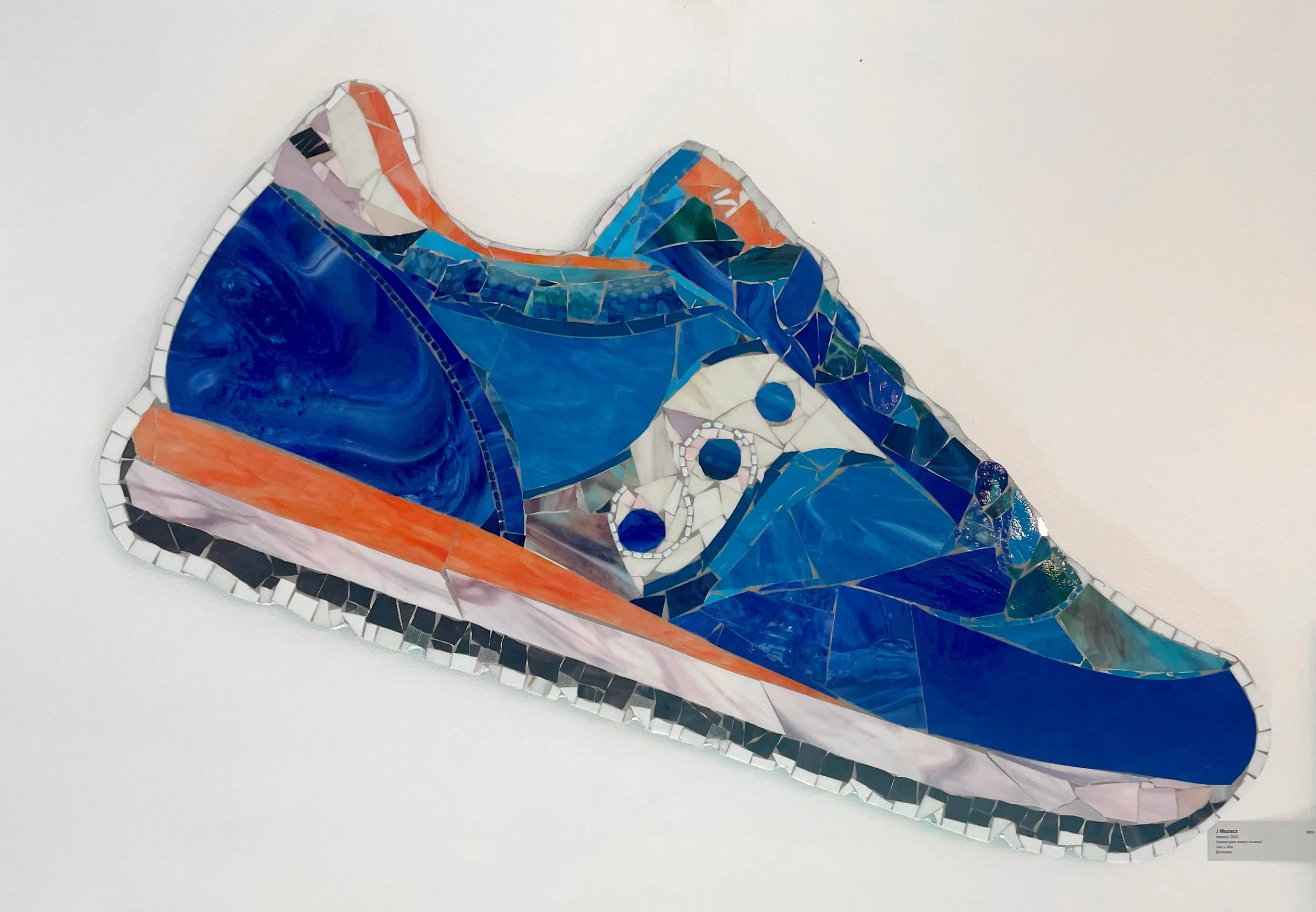 Mosaic artwork of a Saucony sneaker, featuring blue, orange, white, black, and purple tiles, displayed on a white wall.