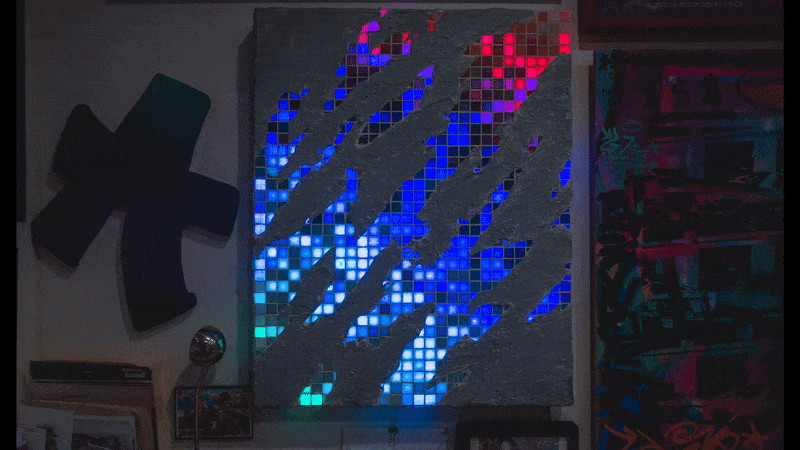 A wall with a large LED light installation displaying colorful, pixelated patterns, accompanied by a black decorative cross and various framed pictures.