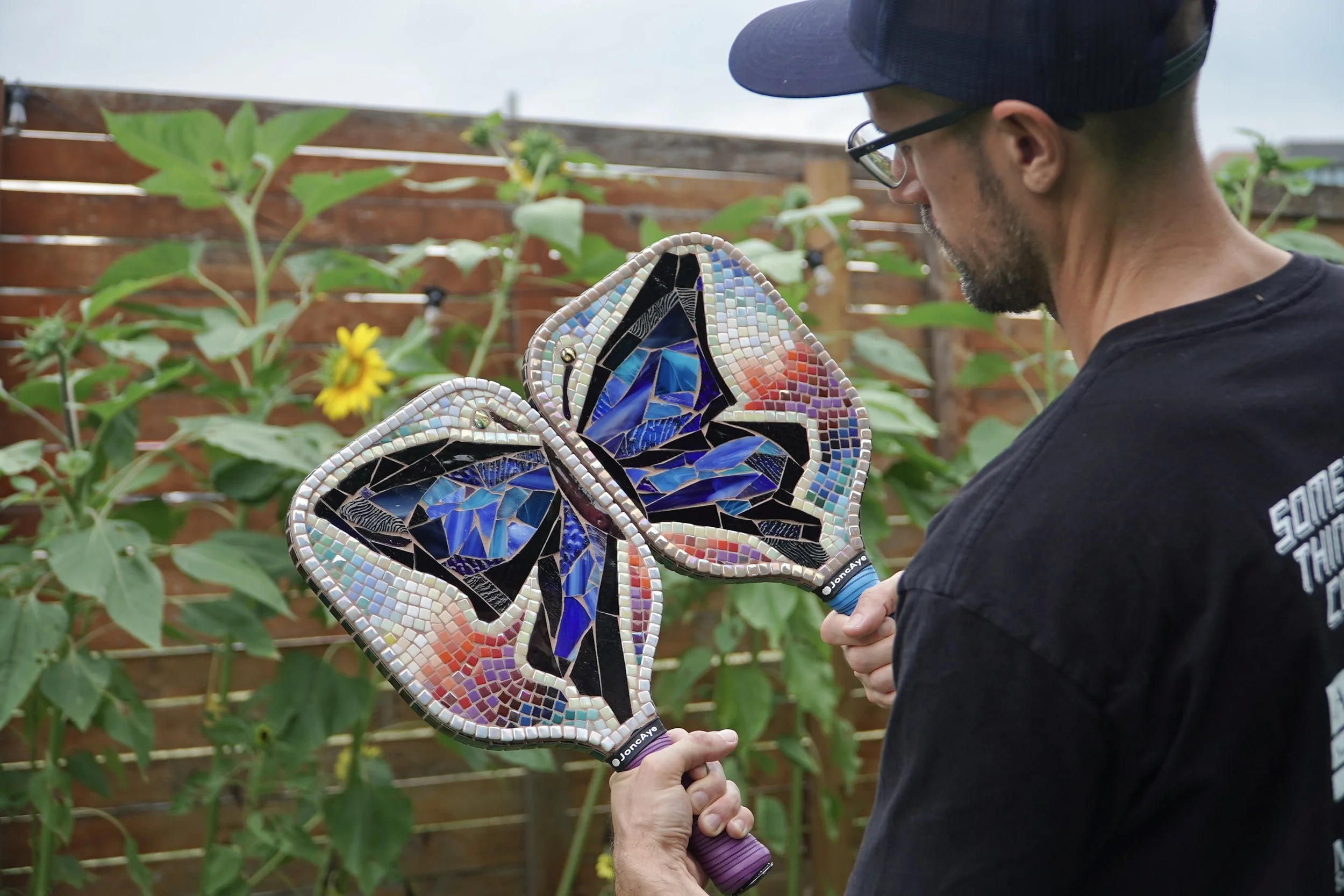A person wearing glasses and a black T-shirt holding two pickleball paddle mosaics each depicting a half of a butterfly.