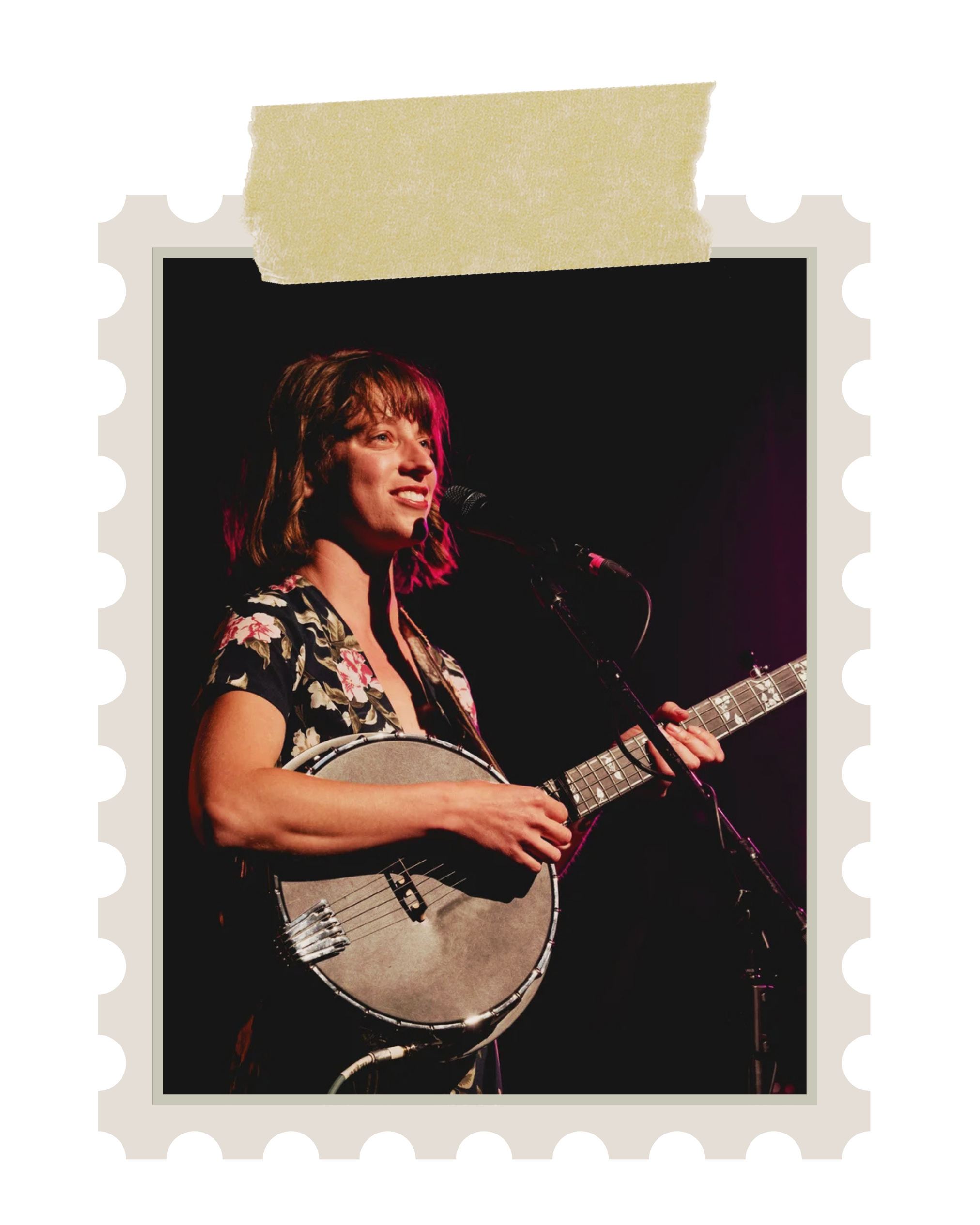 A woman with shoulder-length hair, wearing a black floral top, singing into a microphone while playing an acoustic guitar on stage.