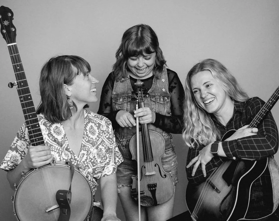 Three women with musical instruments, one holding a banjo, another holding a violin, and the third holding a mandolin, all smiling and interacting in a studio or indoor setting.