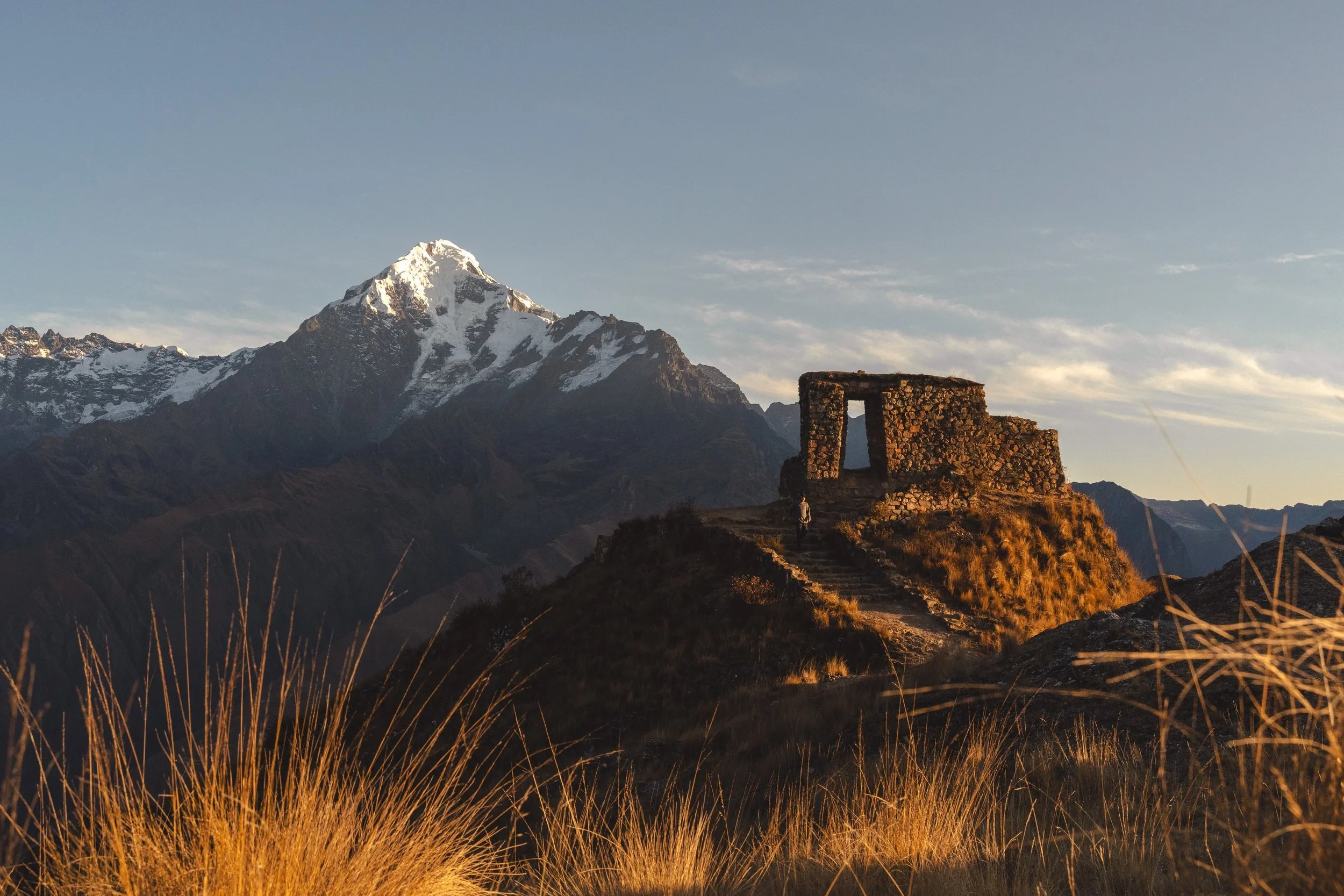 A mountain landscape with Nevado Veronica in the background, an ancient stone structure called Inti Punku on a grassy hill in the foreground, and a clear sky.