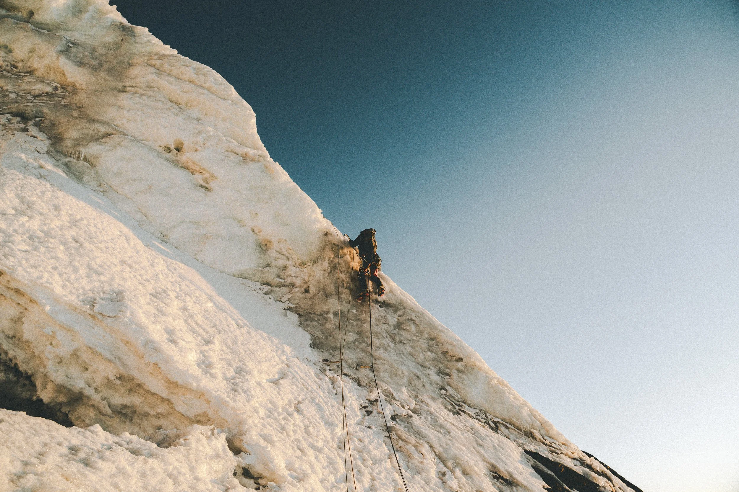 Climber ascending a steep icy glacier with ropes against a clear sky.