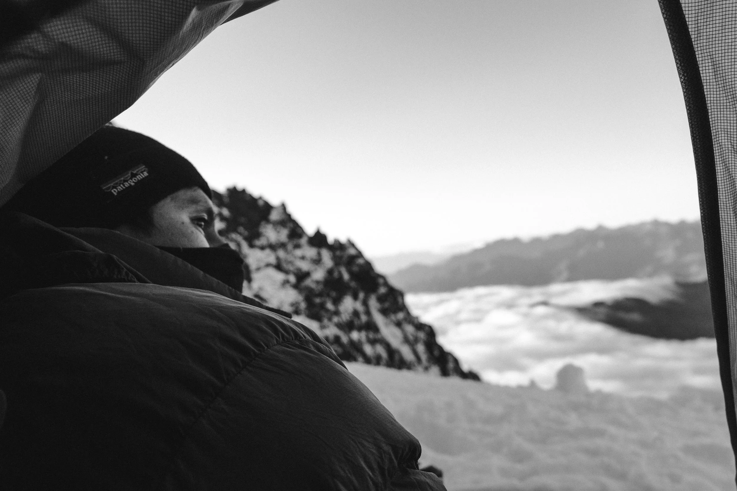 A person in winter gear, wearing a Patagonia beanie, looks out from inside a tent onto snow-covered mountains and clouds.