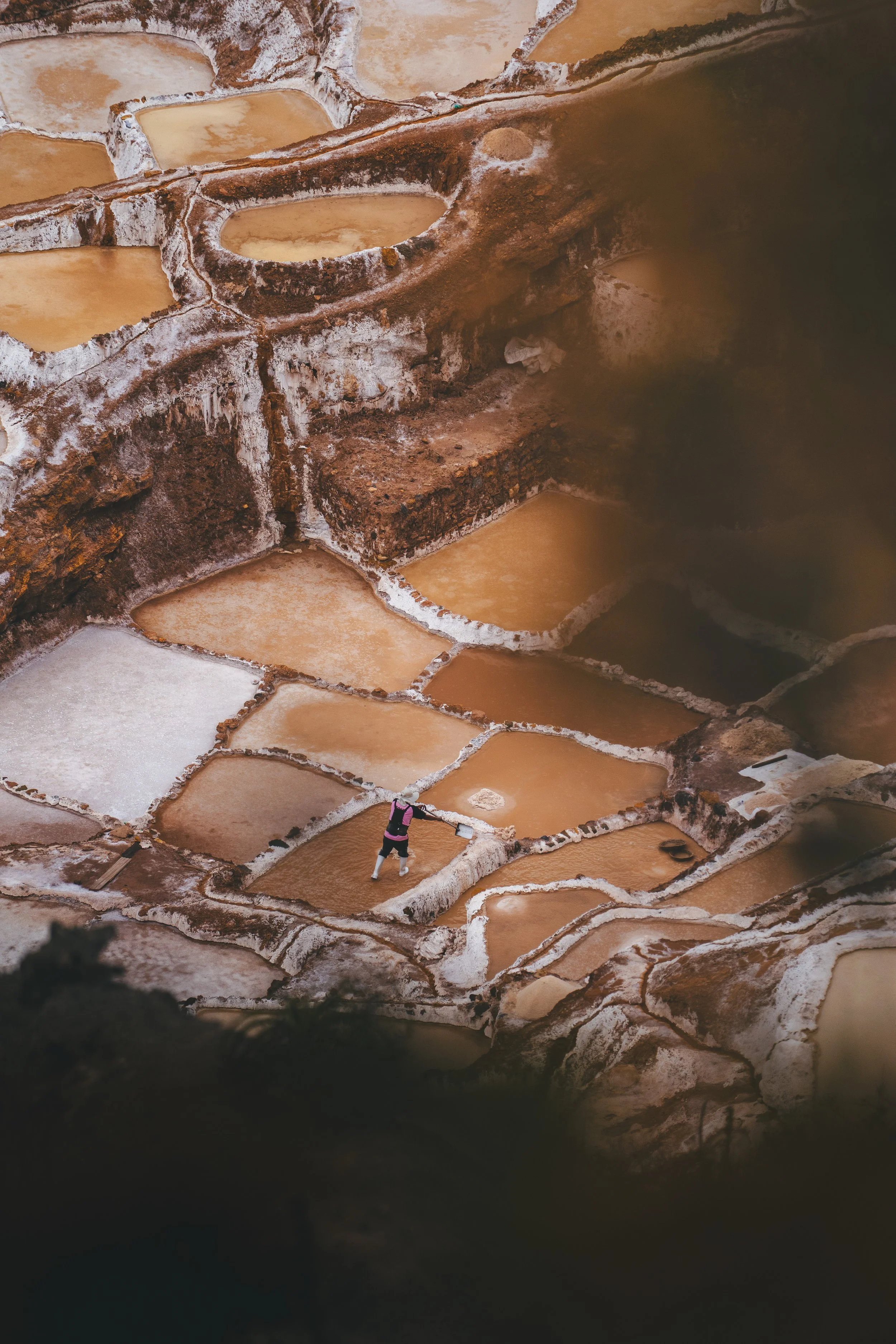 A person working in salt evaporation ponds in the Maras Salt Mines in Urubamba, Peru with multiple shallow pools separated by salt-encrusted edges, mountains in the background.
